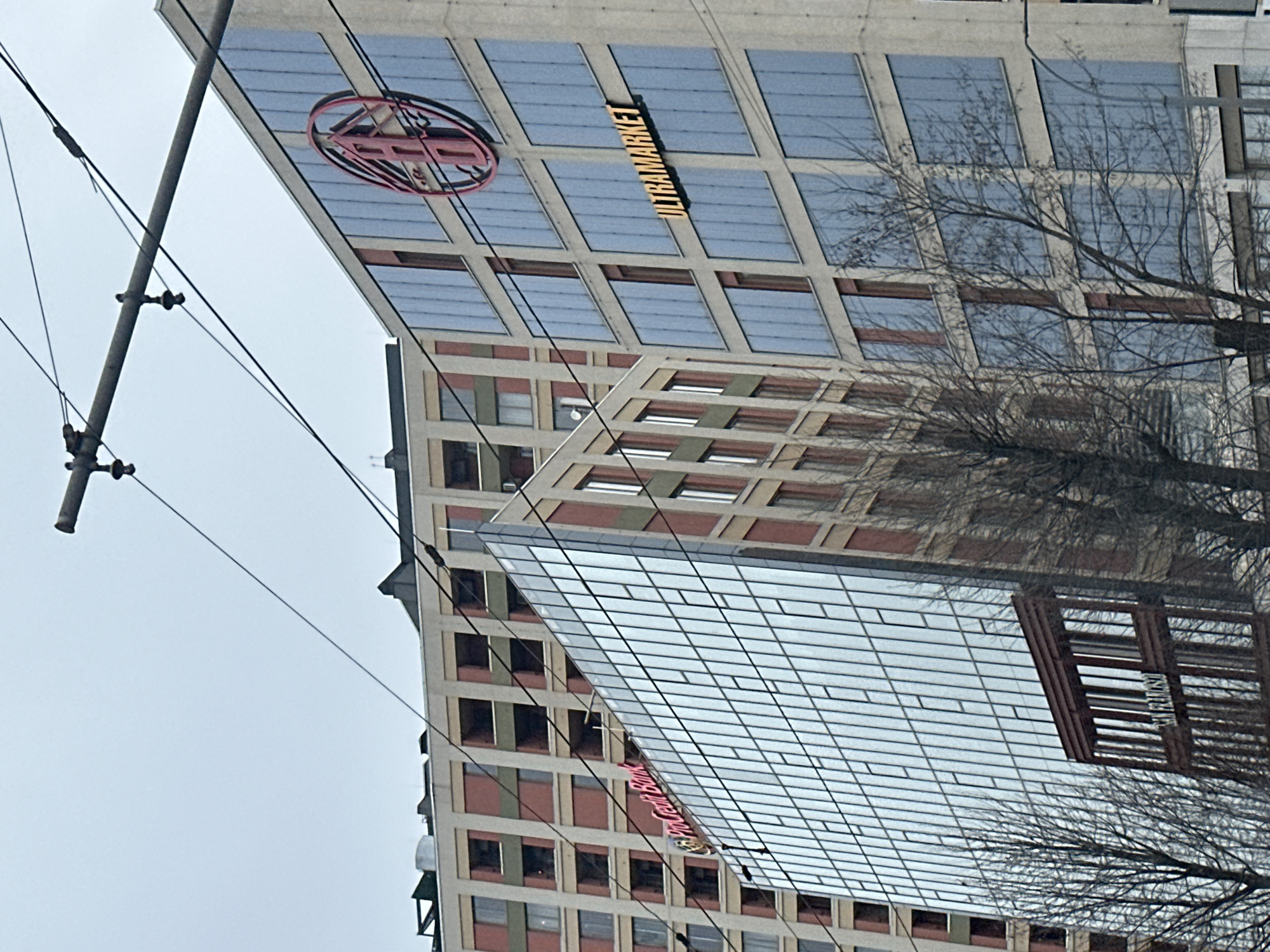 Here's a description of the image:
The image shows a building in Kyiv, Ukraine, specifically the "ULTRAMARKET" building, which is a department store. The building has a grid-like facade with many windows. There is signage indicating the store's name, including a circular logo and the text "ULTRAMARKET." Power lines are visible in the foreground. A bare tree branch is also visible, suggesting it might be winter or early spring. The sky is overcast. The image seems to be taken from a low angle, looking up at the building.