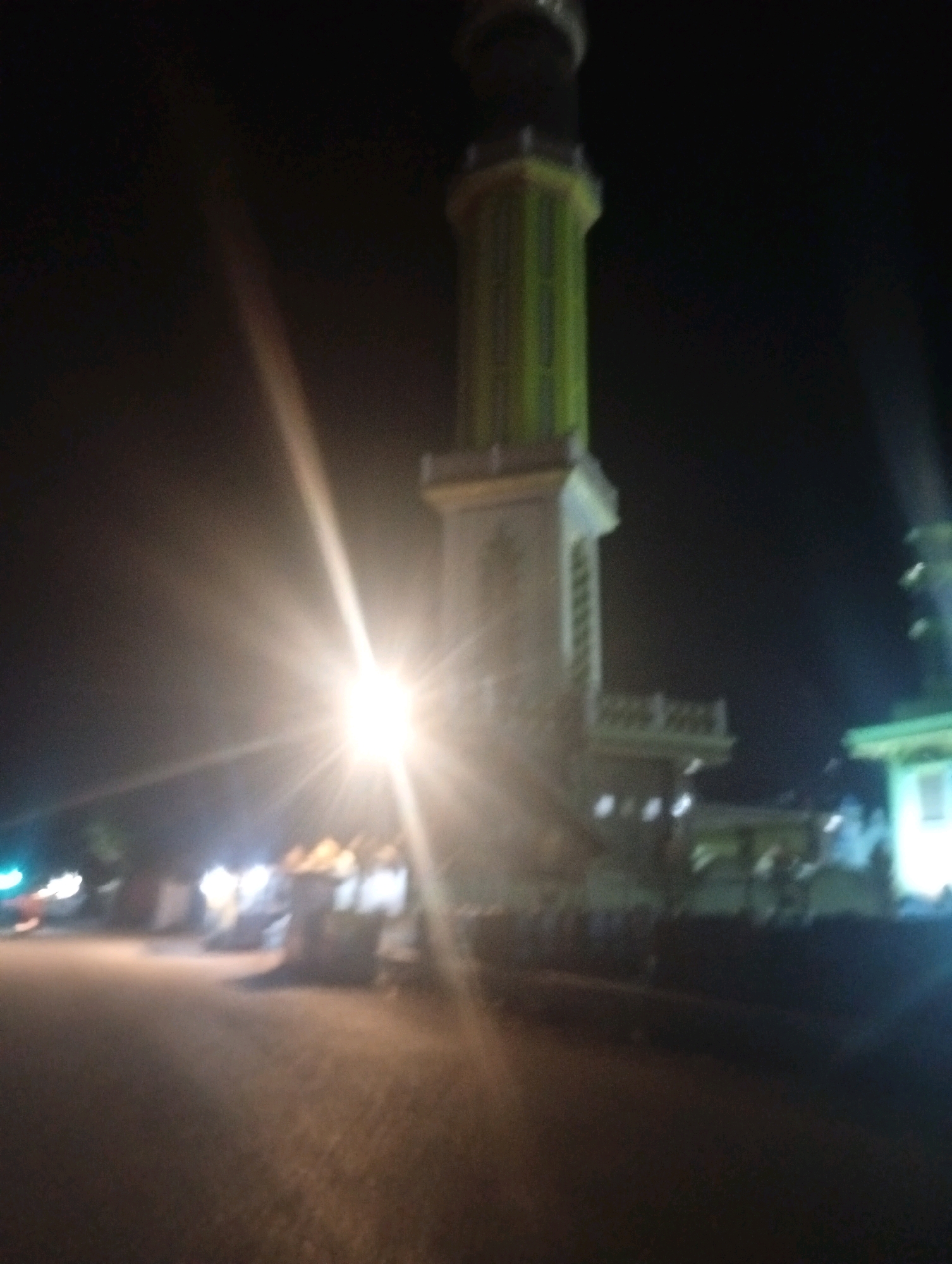 The image shows a minaret at night in Banowan, Indonesia. The minaret is tall and slender, with a light green section near the top. There is a bright light source in the foreground, which is causing lens flare and making the image blurry. In the background, there are other buildings and lights, as well as what appear to be people gathered. The ground in the foreground is dark and appears to be a road or paved area. The overall scene suggests a public gathering or event taking place at night.