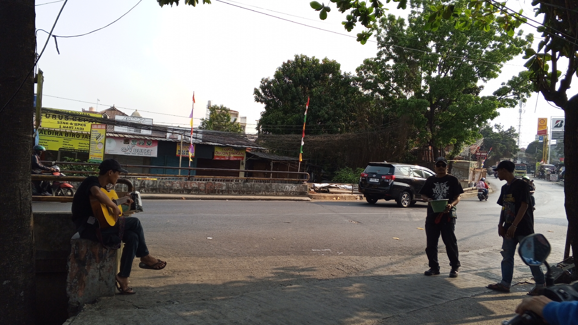 In Bekasi, Indonesia, a street musician with a yellow guitar sits on a concrete block on the side of a road, facing left. He wears a black t-shirt, dark jeans, and flip-flops. Two men stand on the sidewalk to his right. One, wearing a black t-shirt, dark pants, and sunglasses, holds a green bucket and appears to be a street performer or collector. The other, in a black t-shirt and ripped jeans, stands facing the road. A black SUV is parked on the opposite side of the road, with other vehicles and motorcycles visible in the background. Buildings and trees line the street. The scene is captured during daylight.