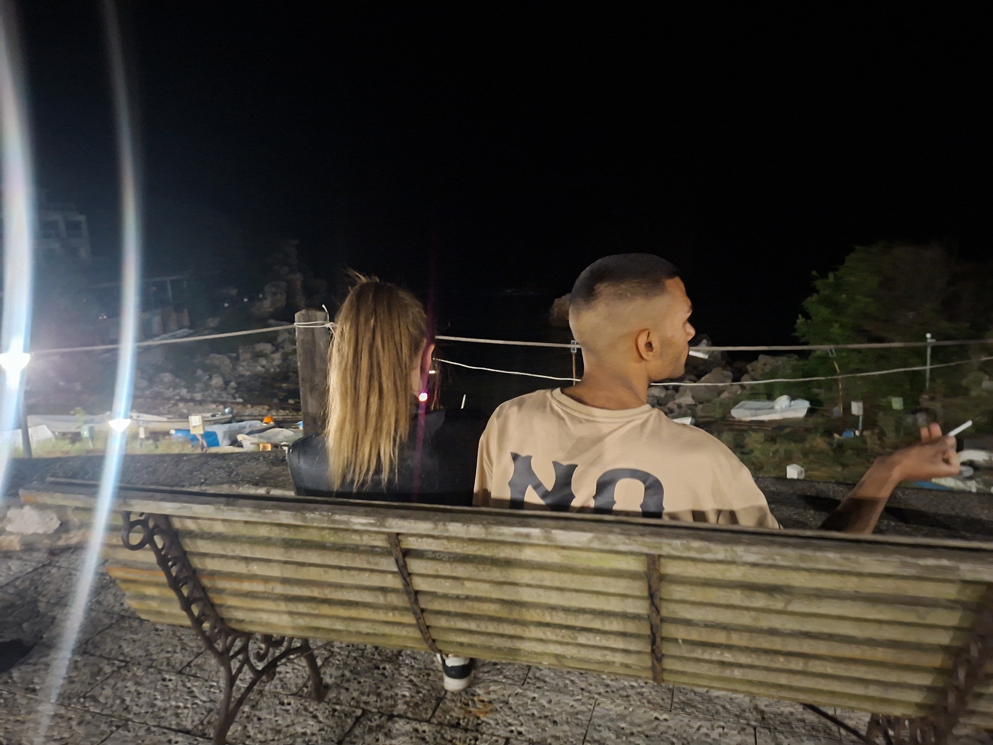 In the city of Tyulenovo, Bulgaria, two individuals are seated on a weathered wooden bench at night. The person on the left, viewed from behind, has long, blonde hair. The person on the right, also viewed from behind, has a closely cropped haircut and is wearing a tan t-shirt with the letters "NO" in a dark, bold font. This individual is holding a lit cigarette in their right hand, with the hand slightly raised. The background is dark, with the faint outline of trees and a body of water visible. The bench is positioned on a textured, pebbled ground. Bright lights in the distance create lens flares across the image.