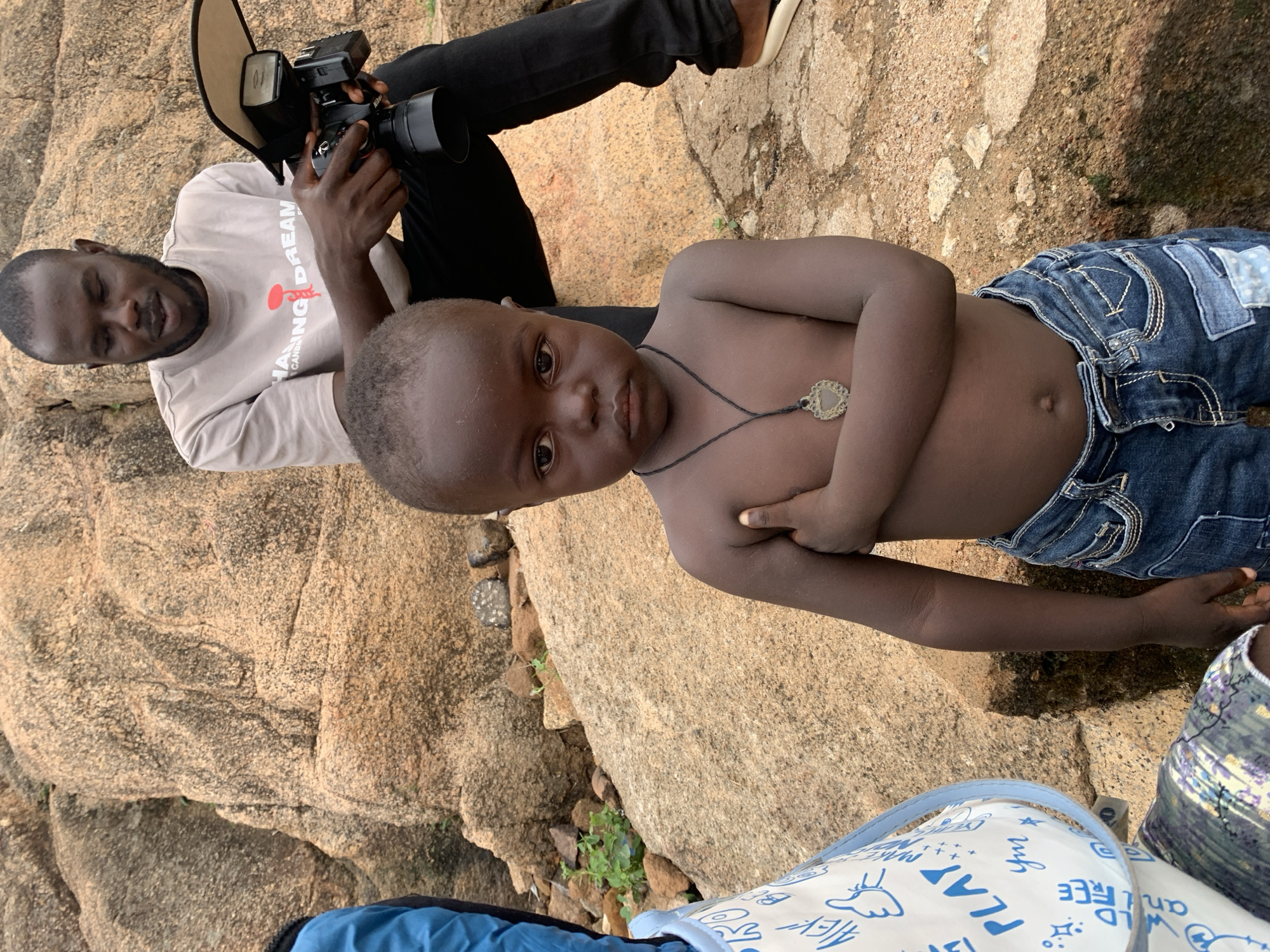 A man is taking a photo of a child in Gura Top, Nigeria. The man is sitting on a rocky surface and holding a camera with a flash. He is wearing a long-sleeved beige shirt with writing on it and black pants. The child is shirtless with blue jeans, wearing a necklace, and posing for the photo. They are also sitting on the rocky surface. A backpack or bag with drawings and writing on it is visible in the foreground.