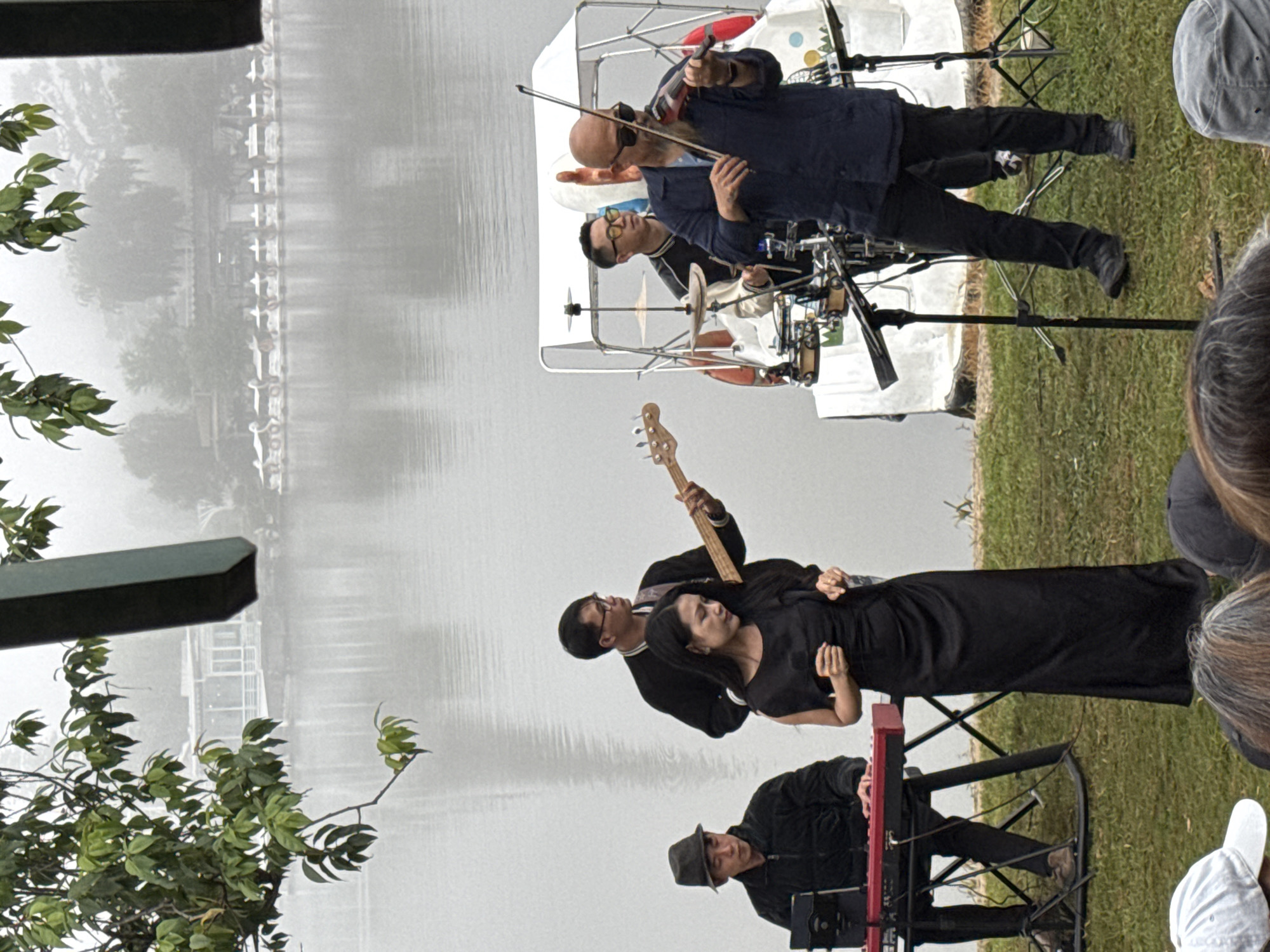 An outdoor performance is taking place by a lake in Da Lat, Vietnam, on a foggy day. A band of four musicians is on stage, playing a mix of instruments. A keyboardist sits on the left, a guitarist in the center, a drummer behind them, and a violinist on the right. A female singer stands in front of the guitarist, holding a microphone and wearing a black dress. The band is on a platform, possibly a boat, decorated with colorful accents. In the background, several swan-shaped boats are docked by the lake, and a row of buildings can be seen across the water. The foreground shows a grassy area where an audience is gathered, with several people wearing hats. The overall atmosphere is serene and artistic, with the fog adding a touch of mystique to the scene.