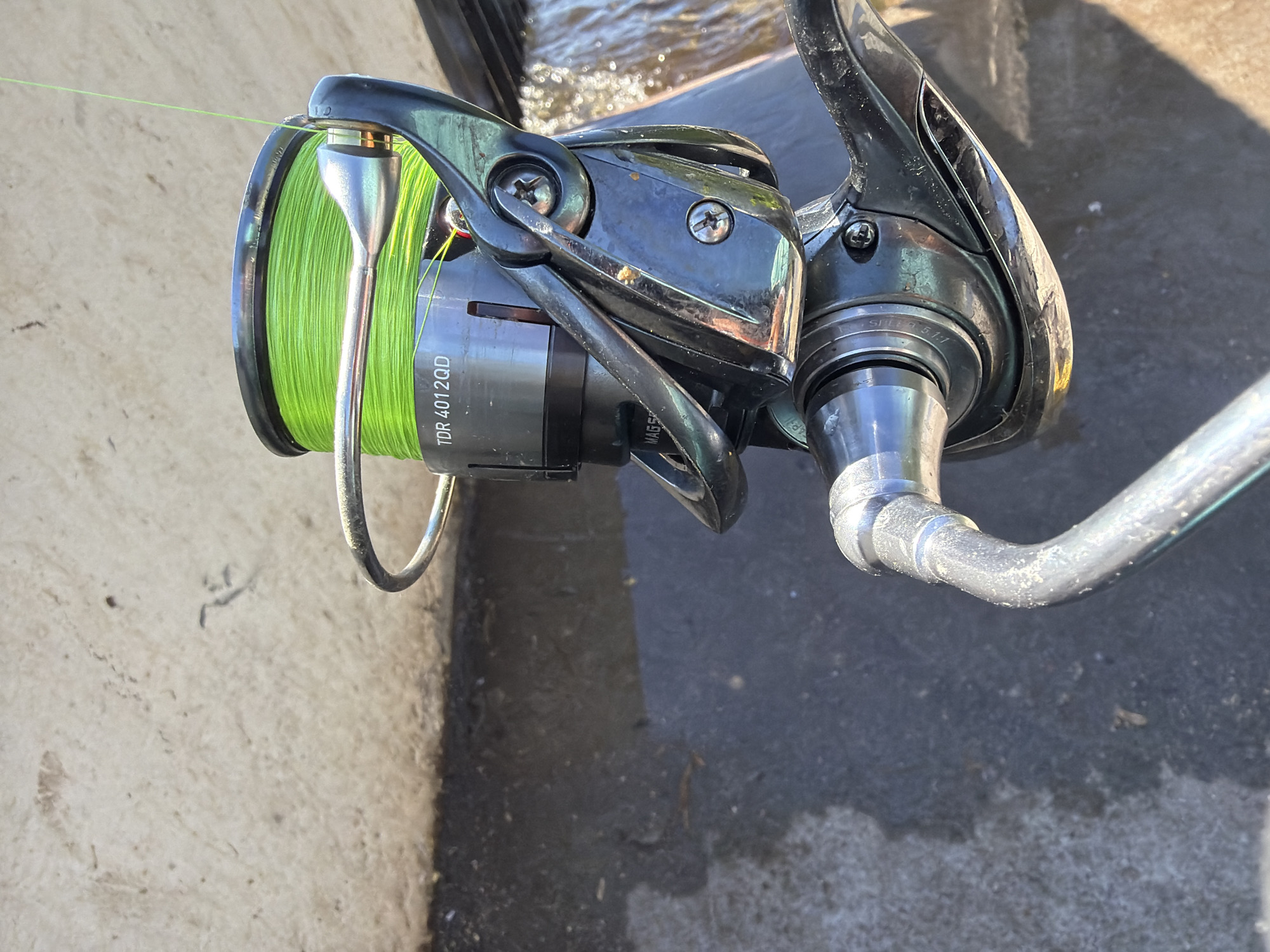 A close-up, high-angle shot captures a fishing reel with bright green line. The reel, which appears to be made of dark metal with silver accents, is positioned over a dark, wet surface with some debris. A portion of a lighter-colored, textured surface is visible in the upper left. The background is indistinct and appears to be outdoors, possibly near a body of water. There are no people or animals visible in the image, and no discernible indicators of the specific location within Chaikovsky, Russia.