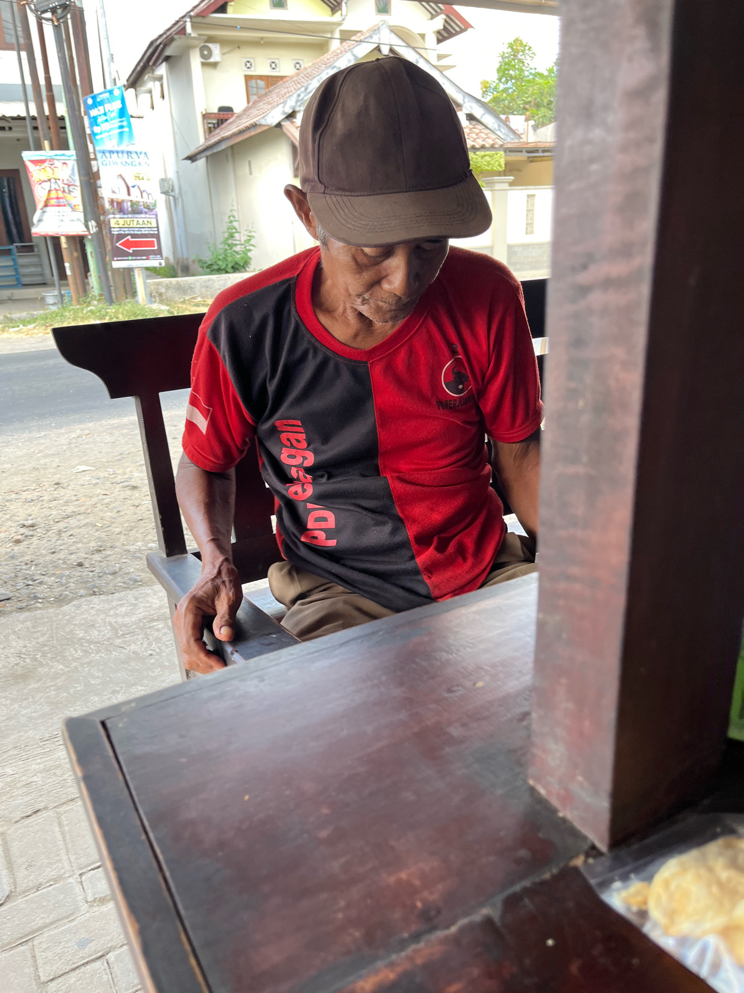 An elderly man wearing a brown baseball cap and a red and black t-shirt with "PD Ega" written vertically on the front is sitting on a dark wooden bench in what appears to be Banguntapan, Indonesia. He is looking down, and his right hand is resting on the bench. In the background, there is a building with a tiled roof, some vegetation, and a street with a sidewalk. There are also several signs visible on poles, one of which advertises "APURYA GIWANGAN" with an arrow pointing left and the text "4 JUTAAN." A portion of a dark wooden table is in the foreground, and a clear plastic bag containing what looks like food is visible on the table. The lighting suggests it is daytime.