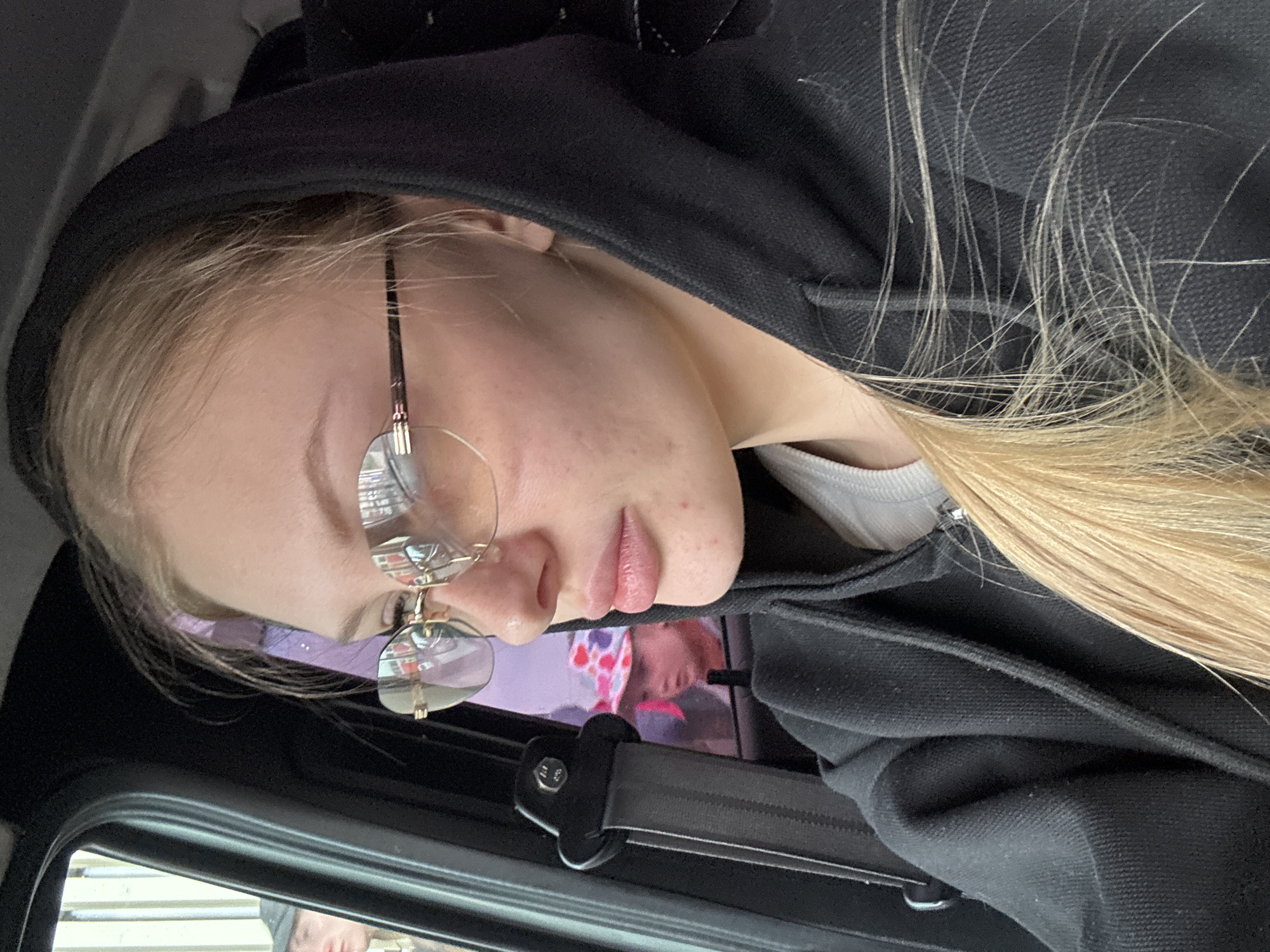 A close-up, angled photograph depicts a fair-skinned person wearing glasses and a black hooded sweatshirt. The person has blond hair and is positioned near a car window with the seatbelt visible. A reflection in the window suggests the location is Perm, Russia, based on the reflected image of someone in front of a building with visible street details.