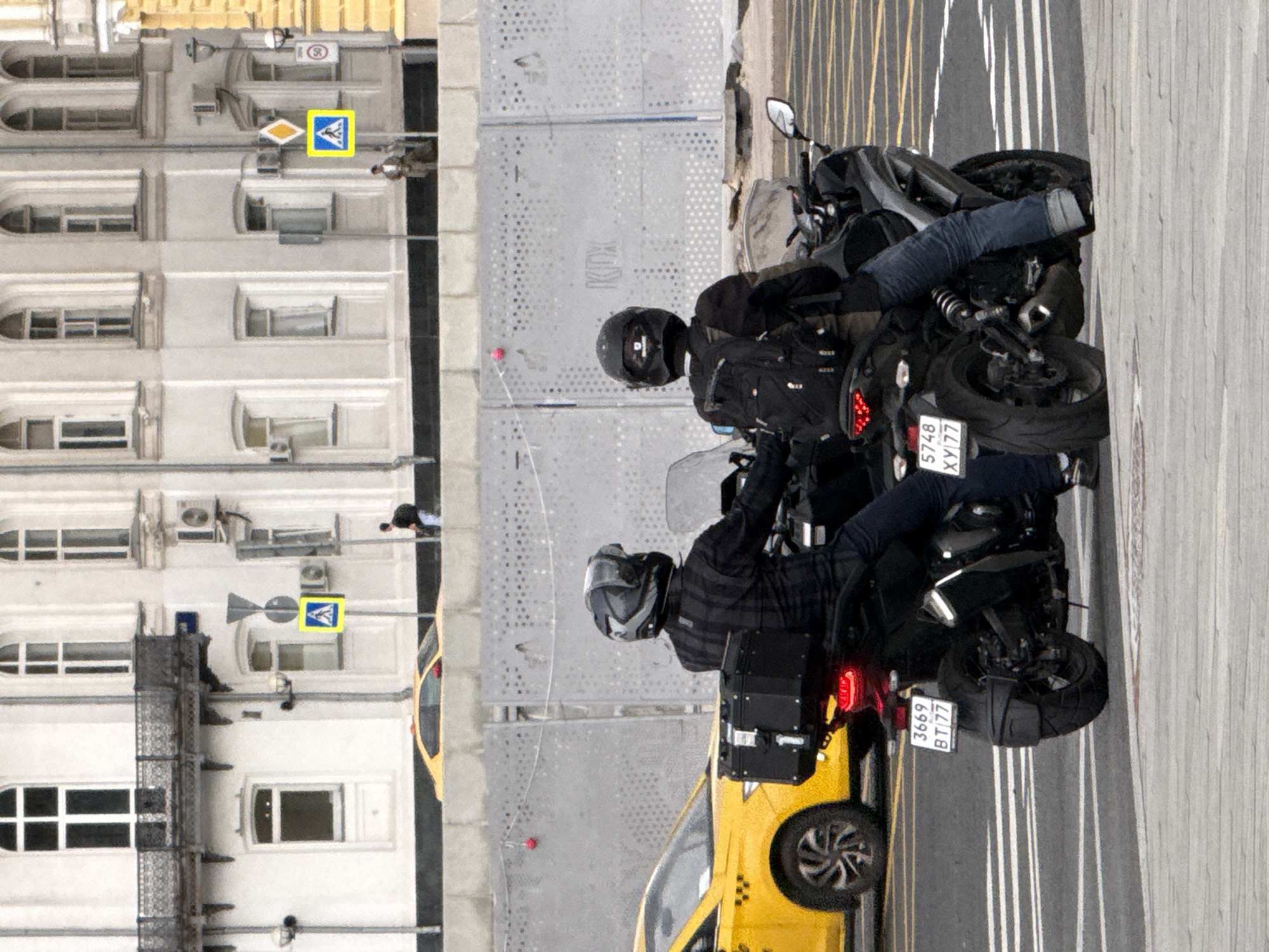 The image depicts a street scene in Moscow, Russia, with two individuals on motorcycles, viewed from behind. The motorcycles are positioned side-by-side, with the rider on the left wearing a plaid shirt and the rider on the right wearing a black jacket and backpack. Both riders have helmets on. In the background, a yellow taxi is visible to the left of the motorcycles. Further in the background, a light-colored building with arched windows and balconies is present, with various signs and traffic signals on poles. A gray barrier runs across the mid-ground of the image. The road surface is asphalt with white lane markings, and the foreground shows a concrete sidewalk or pedestrian area.