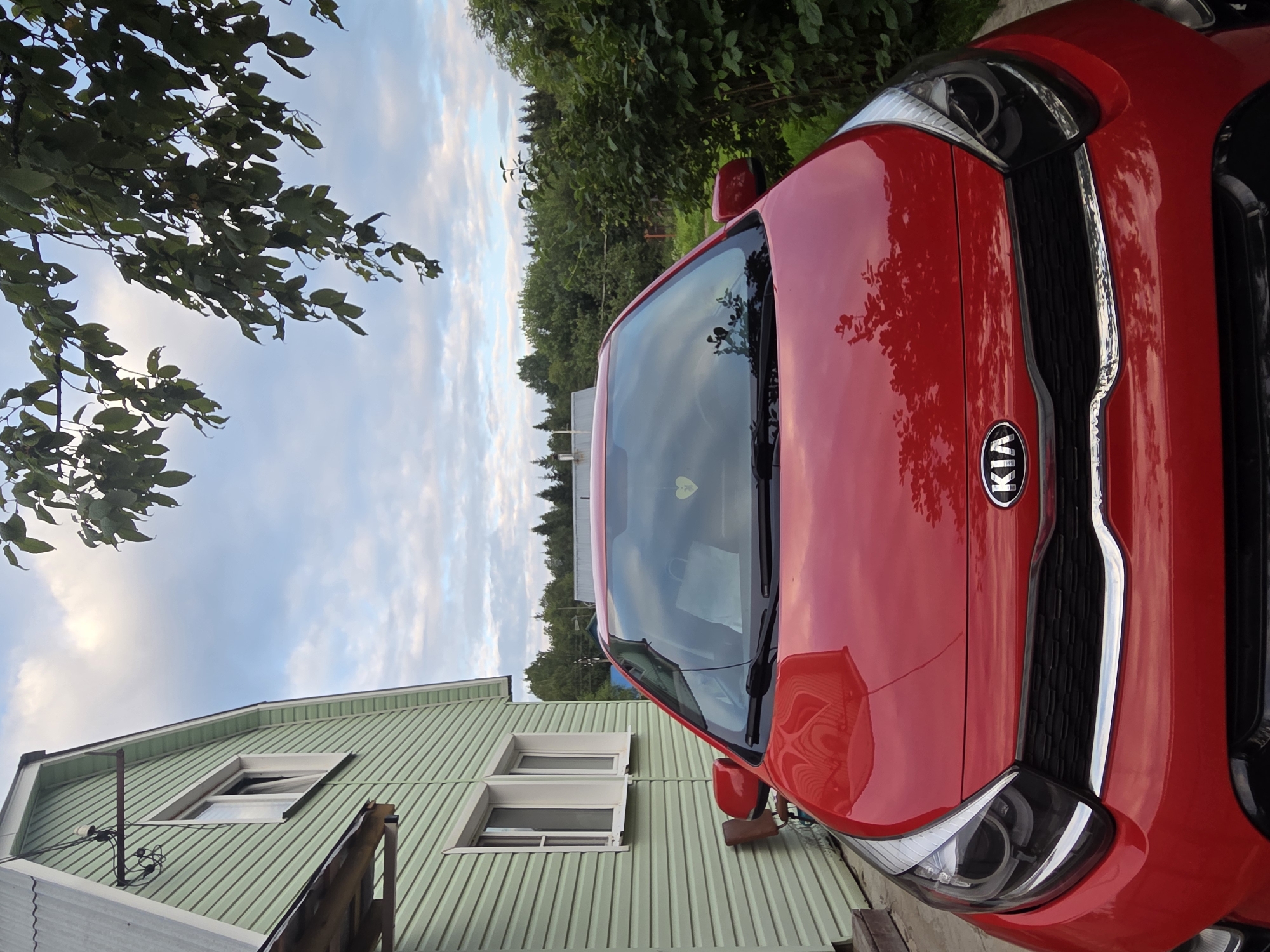 A red KIA car is parked in front of a light green, two-story house in Frolovo, Russia. The house has a metal roof and white window frames. Trees and bushes surround the property, and a cloudy sky is visible above. A branch with green leaves hangs over the car from the top right of the frame.