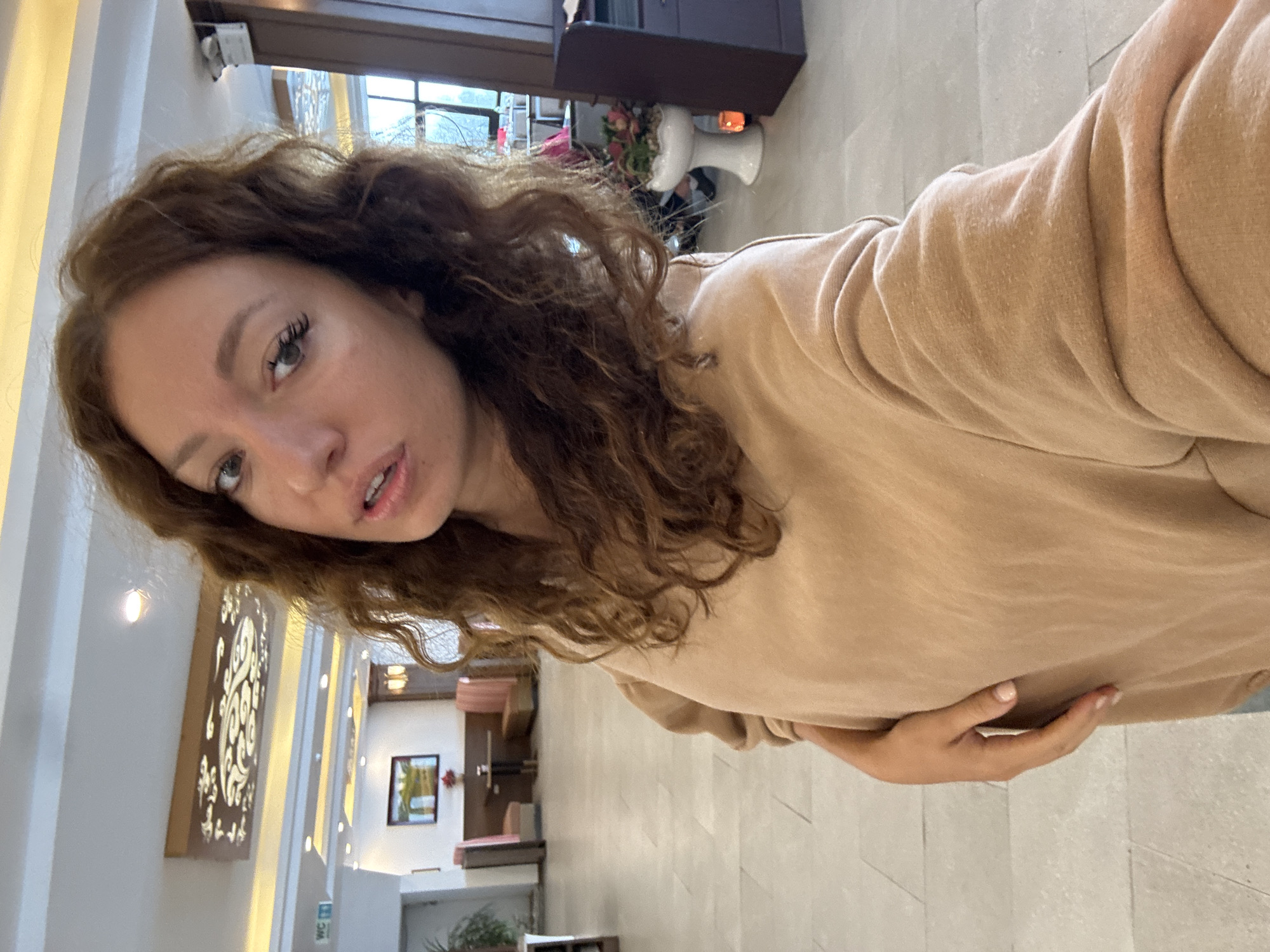 A woman with curly brown hair and fair skin is taking a selfie. She is wearing a beige long-sleeved shirt or sweater. The background shows the interior of a building, likely a hotel or resort, in Binh Lu, Vietnam. Elements in the background include architectural details, decor such as wall art, a doorway, and furniture. A picture frame and plants are also visible.