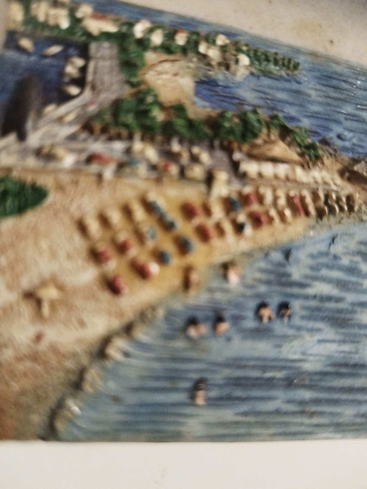 A textured artwork depicts a beach scene. On the beach, small, colorful markers, possibly representing umbrellas or figures, are arranged in rows. People are also seen in the water, indicated by small, dark marks. The water is rendered in shades of blue, suggesting depth. Behind the beach, there are structures possibly houses or buildings, and vegetation. The location is unclear, but it seems to be a beach.