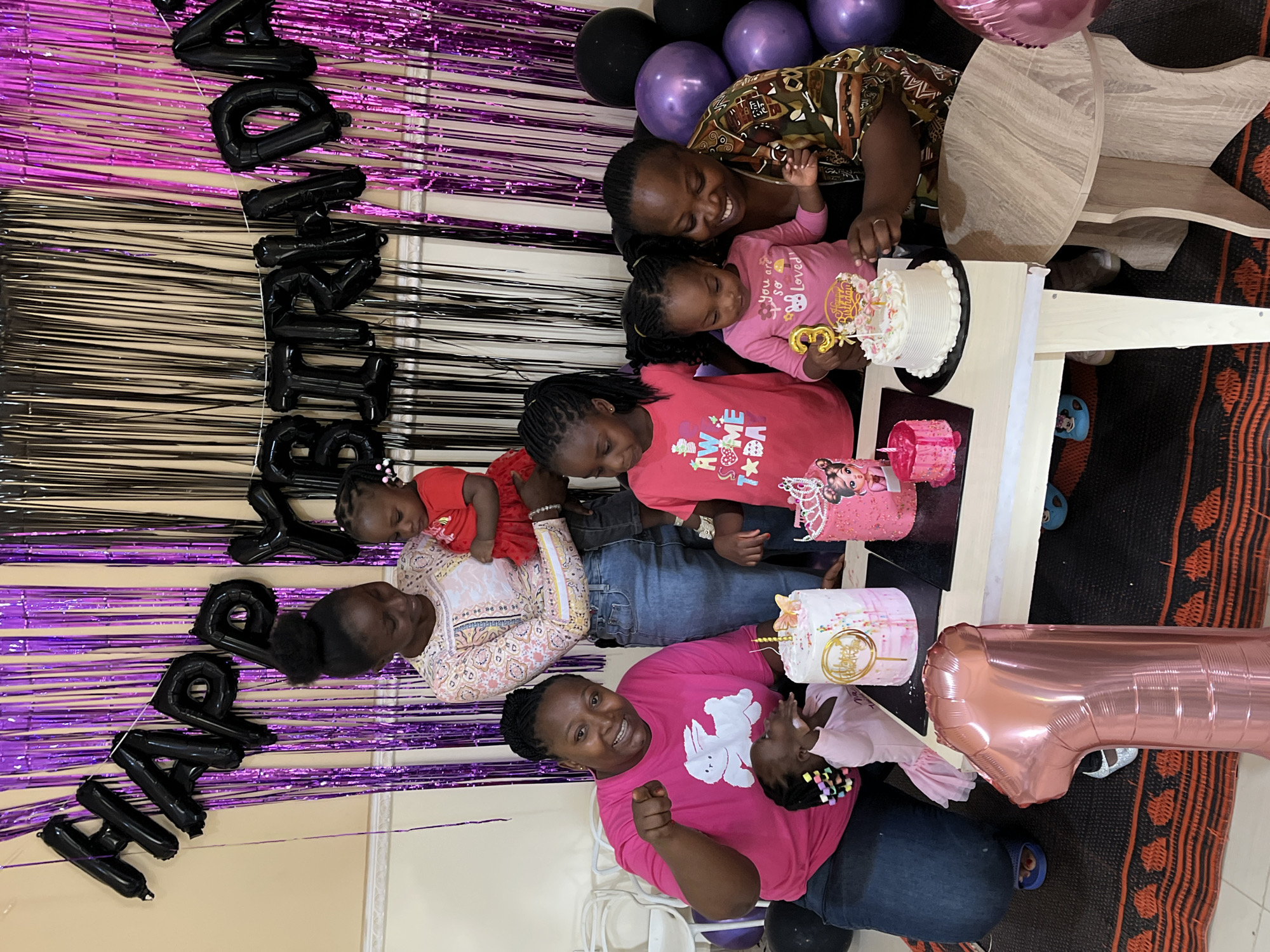 The image depicts an indoor birthday celebration in Kuru Karama, Nigeria, featuring seven individuals: three adult females and four female children, positioned around a light-colored rectangular table. The background is decorated with alternating black and purple metallic fringe curtains, with black foil balloon letters spelling "HAPPY YBIRHDA" (likely "HAPPY BIRTHDAY" with some letters off-frame). Additional black and purple spherical balloons are visible in the upper right background.
On the left, an adult female with braided hair, wearing a pink t-shirt with a white rabbit graphic, blue jeans, and blue slide sandals, kneels. She points towards the viewer with her right hand and smiles. A young female child in a light pink ruffled dress with colorful beaded braids kneels beside her, reaching towards the table. In the center, a standing adult female with dark hair styled in an updo, wearing a patterned long-sleeved blouse and blue jeans, holds a baby in a red dress. Both are looking downwards. To their right, a young female child in a pink t-shirt with "AWESOME" text and stars looks towards the cakes.
In the center-right, a young female child with dark braided pigtails, wearing a light pink long-sleeved t-shirt with "You are so Loved!" text, looks at a white cake with a golden numeral "3" candle/topper, indicating a third birthday celebration. An adult female with dark braided hair, wearing a dark patterned top, kneels next to this child, smiling at her and placing her hand near the cake.
The table displays four cakes: a tall, round white cake with pink frosting accents, sprinkles, a gold "Happy Birthday" topper, a yellow butterfly, and a golden candle; the smaller white cake with the "3" topper and pink floral decorations; a pink-frosted cake with a "Baby Boss" themed character and a silver crown topper; and a very small pink cake with a single candle. On the floor in the immediate foreground, a large rose-gold foil balloon shaped as the numeral "1" stands vertically. The floor is covered by a woven mat with a repeating orange, black, and beige pattern. White plastic chairs are partially visible in the left background.