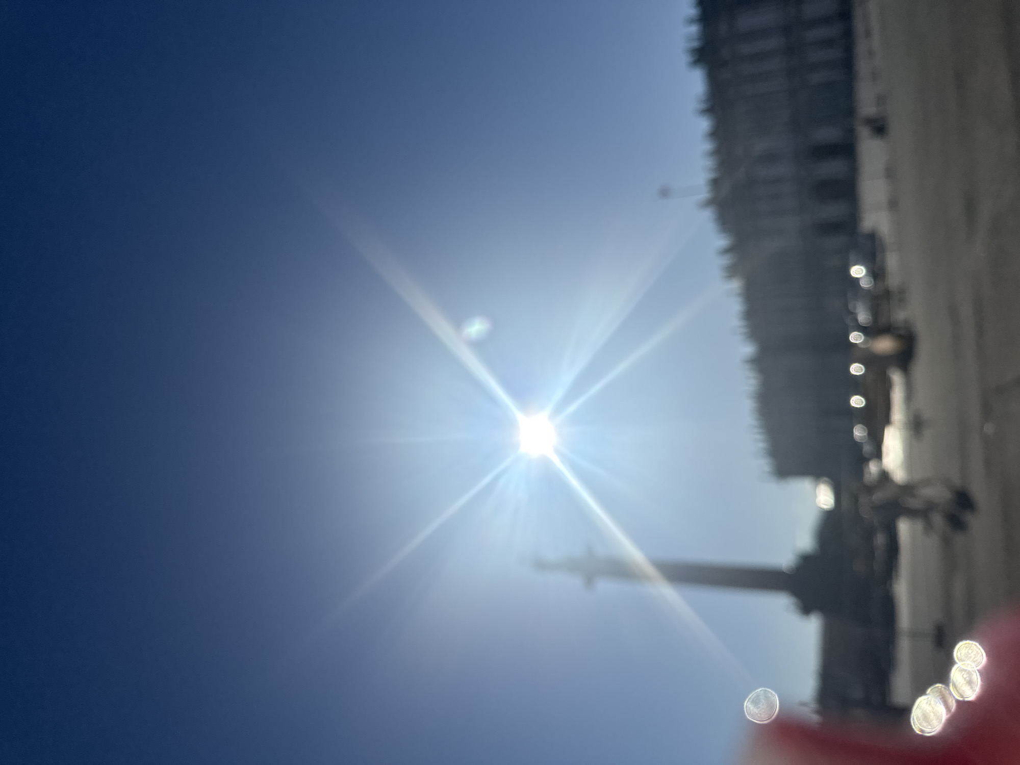 The image depicts a bright, sunny day in Palace Square, Saint Petersburg, Russia. The sun is prominently in the sky, creating a starburst effect and lens flares that obscure some details. In the background, the historic buildings of the General Staff Building and the Winter Palace are visible, though blurred by the sunlight and camera focus. A tall column, likely the Alexander Column, stands to the left, also softened by the glare. Several indistinct figures and what appear to be vehicles are present in the square, suggesting a public space with activity. The overall impression is of a bright, open plaza bathed in sunlight.
