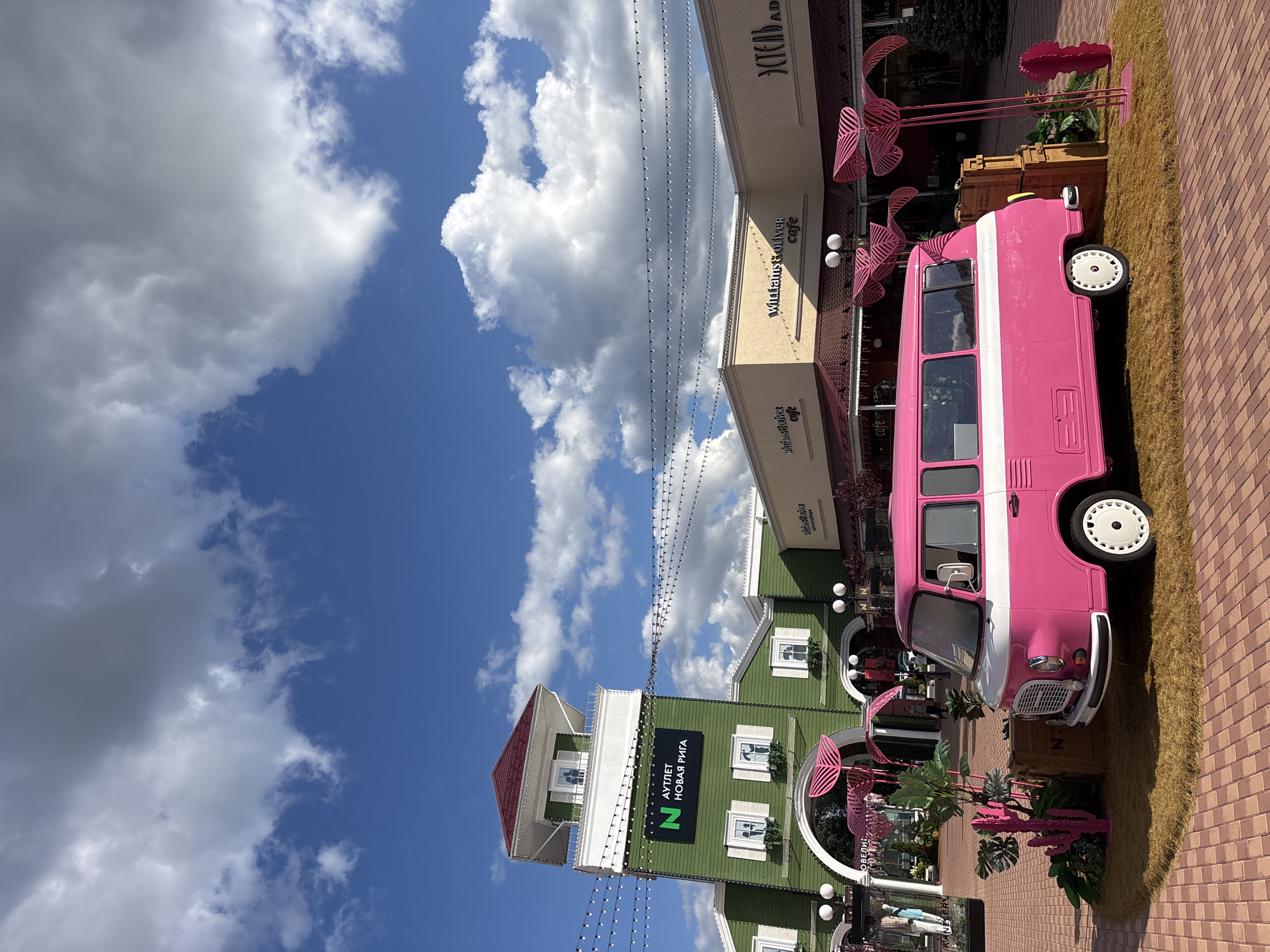 A bright pink and white vintage van is parked on a patch of artificial grass, surrounded by pink tropical-themed decorations, in front of a row of shops under a bright blue sky with scattered white clouds. The van is positioned diagonally, facing the viewer's left. To the left of the van, a tall, green building with a red roof and white trim features a large sign with a green 'N' logo and Russian text. Further to the right, beige buildings with red awnings house various shops, including one labeled "Williams & Oliver cafe." The ground is paved with reddish-brown brick. The scene appears to be an outdoor shopping center or an entertainment district. The location is identified as the Novaya Riga Outlet Village in Pokrovskoe, Russia. There are no people clearly visible interacting with the van or the decorations, though some figures are indistinctly present within the shop entrances.