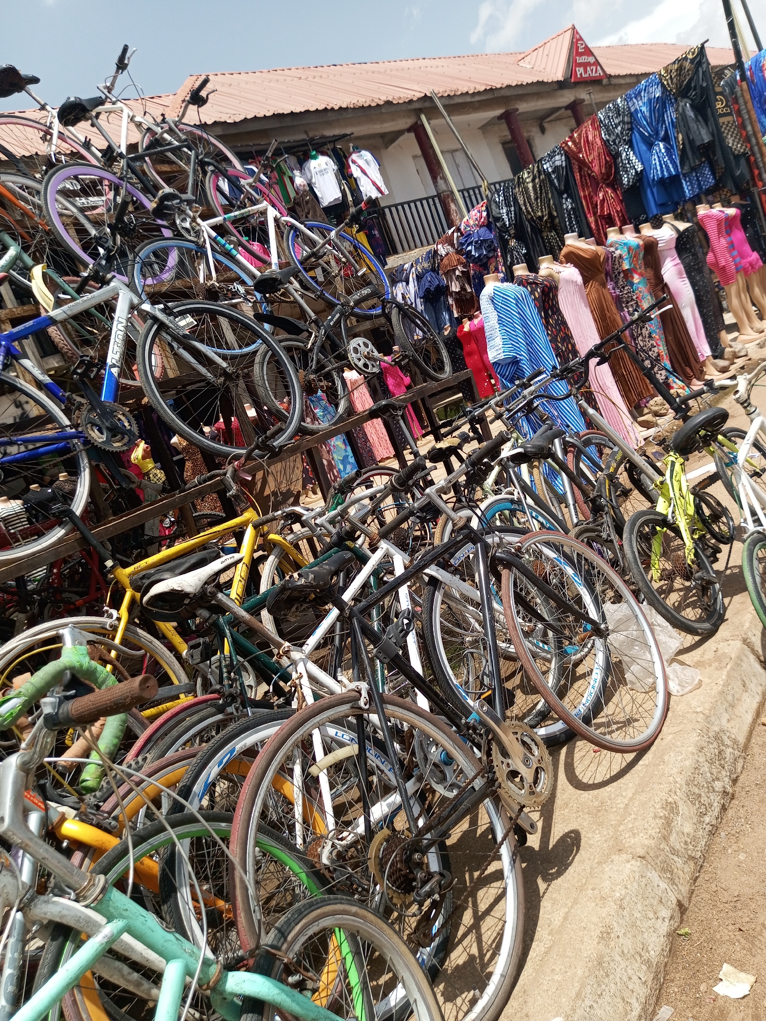 This media file captures a vibrant street scene in Bukuru, Nigeria, likely during the daytime given the bright sunlight and clear sky. The foreground is dominated by a dense collection of bicycles, piled high on wooden racks and leaning against each other. These bicycles vary in color and style, with some appearing more modern and others older and more worn. Their presence suggests a marketplace or a repair shop where bicycles are being sold or serviced.
Behind the bicycles, a row of shops is visible, characterized by their terracotta-tiled roofs and visible structures. In front of the shops, various garments are displayed on racks and mannequins. These include brightly colored dresses, robes, and what appear to be traditional African attire, indicating a lively retail environment. The mannequins, some partially visible, are dressed in colorful fabrics, adding to the visual richness of the scene.
The ground is a rough, concrete surface, suggesting an outdoor setting, possibly a market street. There are some scattered pieces of paper or plastic on the ground, typical of an urban environment. The overall impression is one of a busy commercial hub, with a strong emphasis on both the bicycle trade and the textile and fashion industry.
A sign with red lettering on a white background is visible above the shops, reading "PLAZA". This offers a specific location cue within Bukuru. The abundance of goods and the organized yet somewhat chaotic arrangement of the bicycles and clothing create a sense of bustling activity and local commerce. The weather appears to be fair and sunny, contributing to the overall brightness of the image. There are no people prominently featured in the image, the focus being on the objects and the setting.