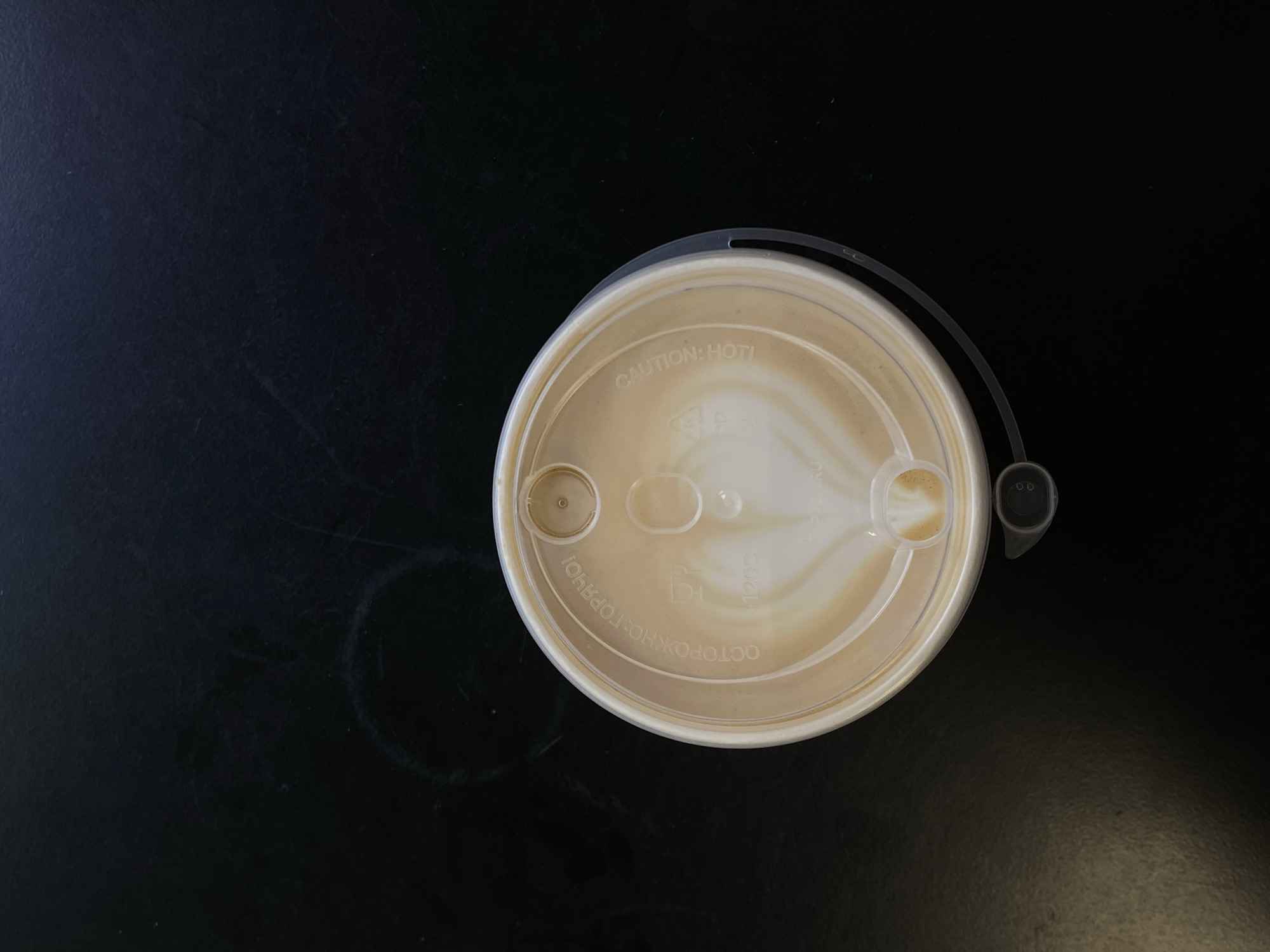 A top-down shot shows a coffee cup lid on a dark surface. The lid is round and off-white, with "CAUTION: HOT!" printed on it. The cup is presumably filled with coffee, visible through the transparent lid. The background is a dark, possibly black, surface, with a faint ring stain towards the left side. There is nothing in the image to indicate a specific location in Saint Petersburg or any other location.