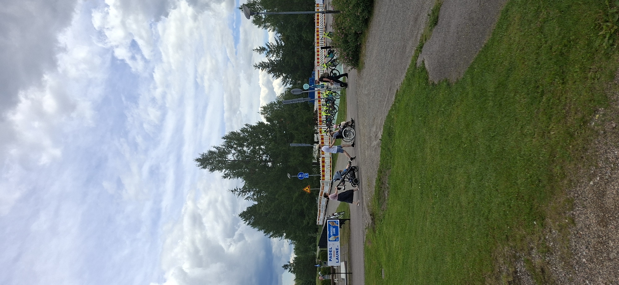 The image shows a scene at a park or recreational area, likely in Lahti, Finland, given the Finnish text on a sign. The sky is overcast with dramatic clouds. In the foreground, there is a well-maintained green lawn. A gravel path curves from the lower right towards the center of the image.
Several people are present and engaged in various activities. A woman in a dark skirt and light top is pushing a stroller with a child. Next to her, a man in a white t-shirt and blue shorts is walking, seemingly accompanying the stroller or the person in the wheelchair. A person in a wheelchair, possibly a child or a smaller adult, is being pushed by someone whose lower body is visible. Further to the right, a person in a dark shirt and dark pants is walking.
In the background, there is a row of white and yellow barricades, suggesting some sort of event or ongoing construction. Behind the barricades, numerous bicycles are parked, many of them yellow, indicating a bike rental or sharing service. Trees, predominantly coniferous, form a dense forest line behind the barricades and bicycles.
On the left side of the image, a sign is visible with Finnish text that appears to read "PADEL LAUNGE" and "VARAA MINIGOLF LAUNE.FI". This confirms the location is likely related to leisure activities, with Padel and Minigolf mentioned, and it strongly suggests a connection to Lahti. Traffic signs, including a pedestrian crossing symbol and a yellow warning triangle, are also visible near the barricades. The overall atmosphere is one of a public space being utilized for recreation under a dynamic sky.