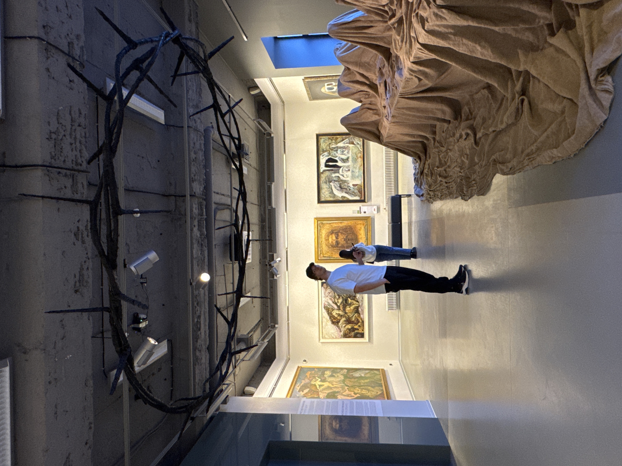 The image displays the interior of an art gallery or museum, captured from a slightly tilted perspective, giving a dynamic view of the space and its exhibits. Two visitors are present, observing the artworks.
On the left side of the frame, a massive, dark, sculptural installation dominates the foreground. It resembles an oversized crown of thorns or a complex arrangement of sharp, spiky branches, possibly crafted from metal, extending from the floor to the ceiling. Contrasting with this is a large, flowing sculpture made of voluminous, light brown fabric draped dramatically from the ceiling or upper wall on the right side, creating deep folds and texture.
Two individuals are engaged in viewing the art. A man wearing a white t-shirt, dark trousers, and sneakers stands in the mid-ground, gazing upwards and towards the paintings on the opposite wall. Further back, another person in a light top and blue jeans is also seen observing the exhibits.
The gallery walls are a neutral light color, showcasing several framed paintings. Notable among them is a large artwork behind the man, featuring abstract or expressive figures in tones of green and brown. Above it, a smaller painting displays the prominent letter "B." Other paintings with more classical or figurative elements are visible along the walls, illuminated by both spotlights and linear ceiling lights. A skylight in the ceiling provides natural light, casting a blue hue into the space. The floor is a smooth, light grey. A display case or information panel is partially visible in the bottom foreground, though any text on it is unreadable. The overall atmosphere suggests a quiet and contemplative art viewing experience within the museum setting in Saint Petersburg, Russia.