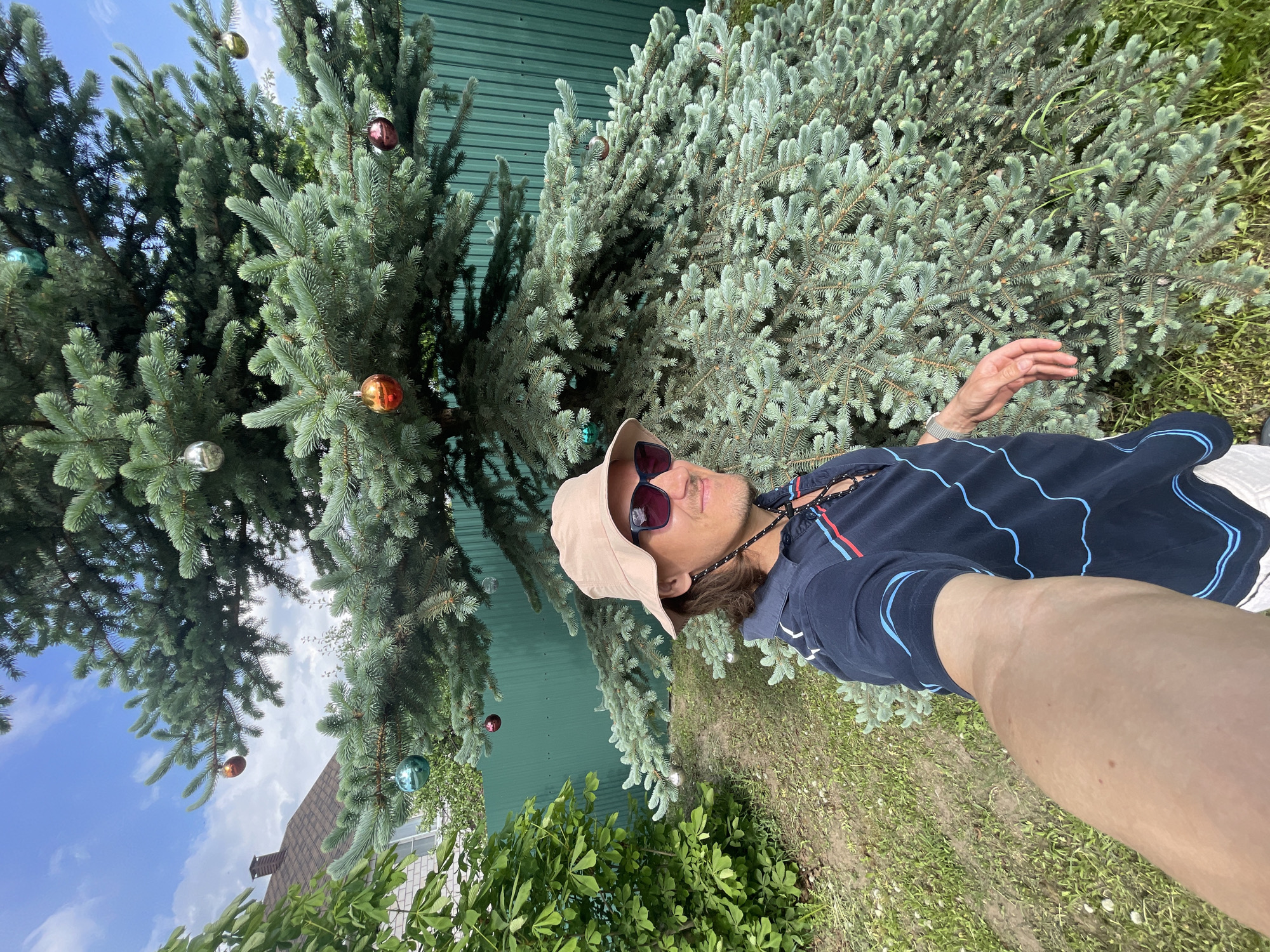 A man wearing a beige bucket hat and sunglasses stands in a yard next to two evergreen trees that are decorated with ornaments. The man is taking a selfie, and the trees and a green corrugated metal fence are in the background. A patch of grass is visible in the foreground. It is daytime. A glimpse of a house with a brown roof can be seen to the left of the evergreen trees. There is no discernible evidence to indicate that this image was taken in Izhevsk, Russia.