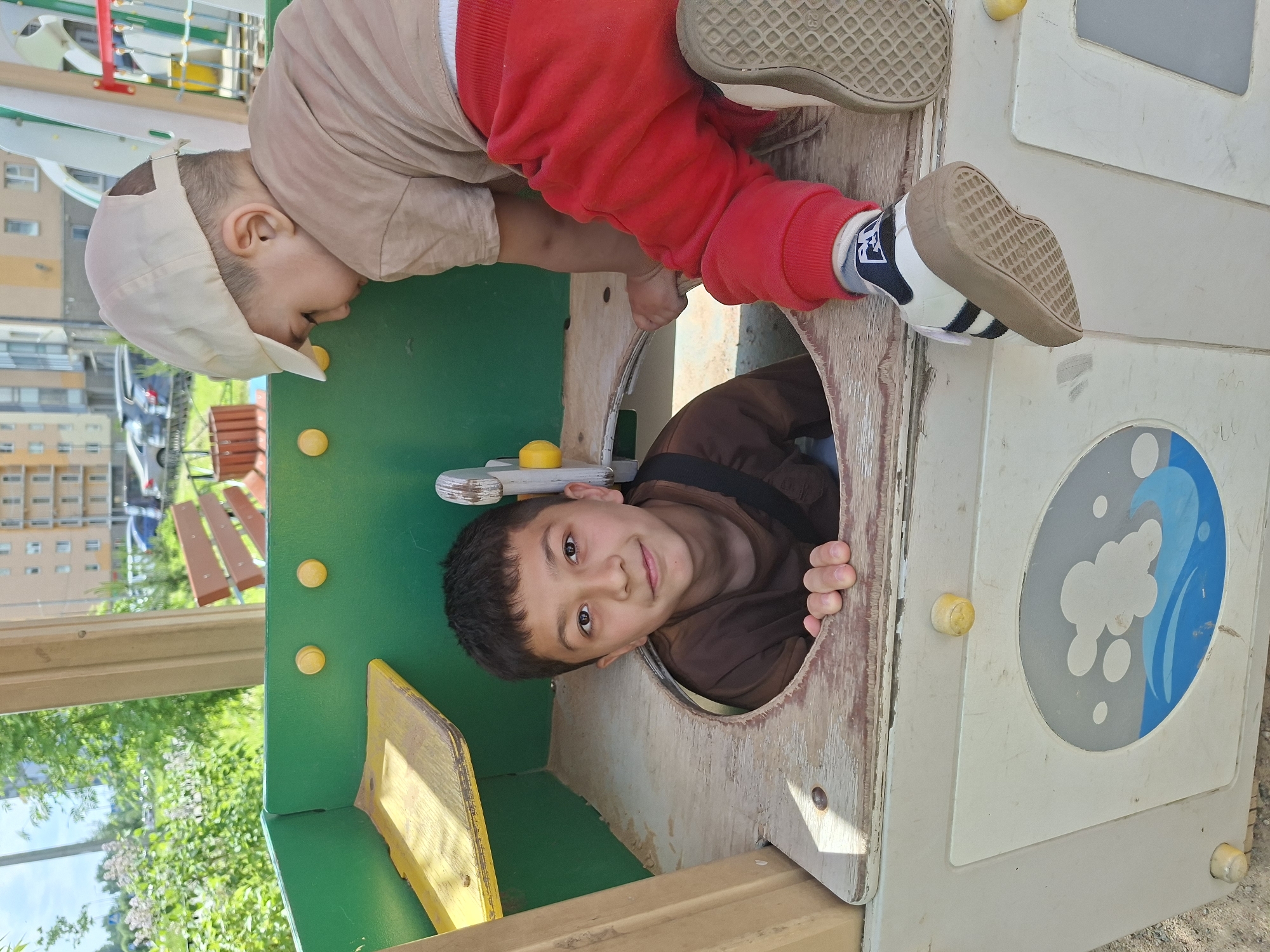 The image captures two children interacting with a playground structure under natural daylight in Unknown, Russia.
In the foreground, a multi-component playground structure, primarily green, light brown, and beige, occupies the lower and central portions of the frame. The materials appear to be faded plastic or painted wood. On the left, a green wall features a small yellow shelf. In the center, an older boy, approximately 8-12 years old, with dark hair and dark eyes, emerges from a circular opening in a light brown panel. He wears a dark brown t-shirt with a visible black strap and looks directly at the camera with a slight smile, his hands resting on the edge of the opening. To his right, a younger boy, approximately 1-3 years old, wearing a light-colored t-shirt, red sweatpants, and a beige baseball cap worn backward, is in the process of climbing onto the structure. His face is turned downwards, focused on his movement, and his left foot, clad in a white sneaker with black stripes and a tan sole, is visible stepping onto a worn wooden surface of the structure. A light beige panel at the bottom right features an oval design depicting blue waves and white cloud-like shapes. Yellow knob-like elements are present on various parts of the playground equipment. The ground beneath the structure appears to be sand or dirt.
The background reveals a residential setting with lush green foliage, including trees and bushes, on the left. On the right, multi-story residential buildings with light-colored facades are visible. Several parked cars can be seen on a street or parking area behind the buildings. The scene is brightly illuminated, indicating a clear, sunny day.