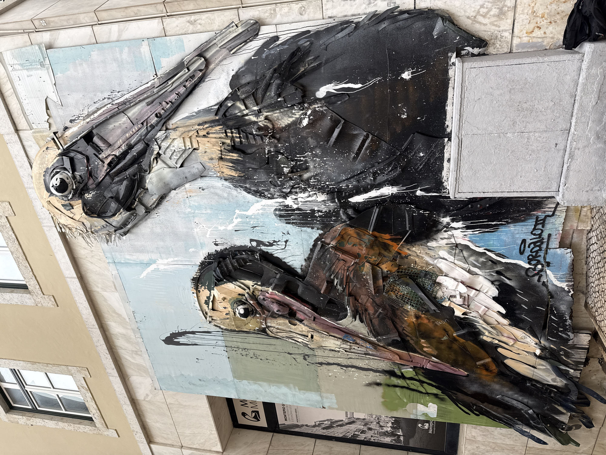 The image shows a close-up, tilted view of a street art mural of two birds located in Lisbon, Portugal. The birds are constructed from various materials, possibly recycled items, and are painted with black, white, brown, and grey hues. They feature distinct eyes and beaks, with one bird positioned above the other. Paint splatters and graffiti are present on the background.
The mural is affixed to the side of a building with a pale yellow facade and windows with black frames. Below the artwork, there is a sign with the words "MONTEPIO G", "MONTEPIO GERAL" and "BOROLO" visible, along with a picture. The building's exterior is finished with marble-like tiles. A black garment hangs at the top right corner of the frame.