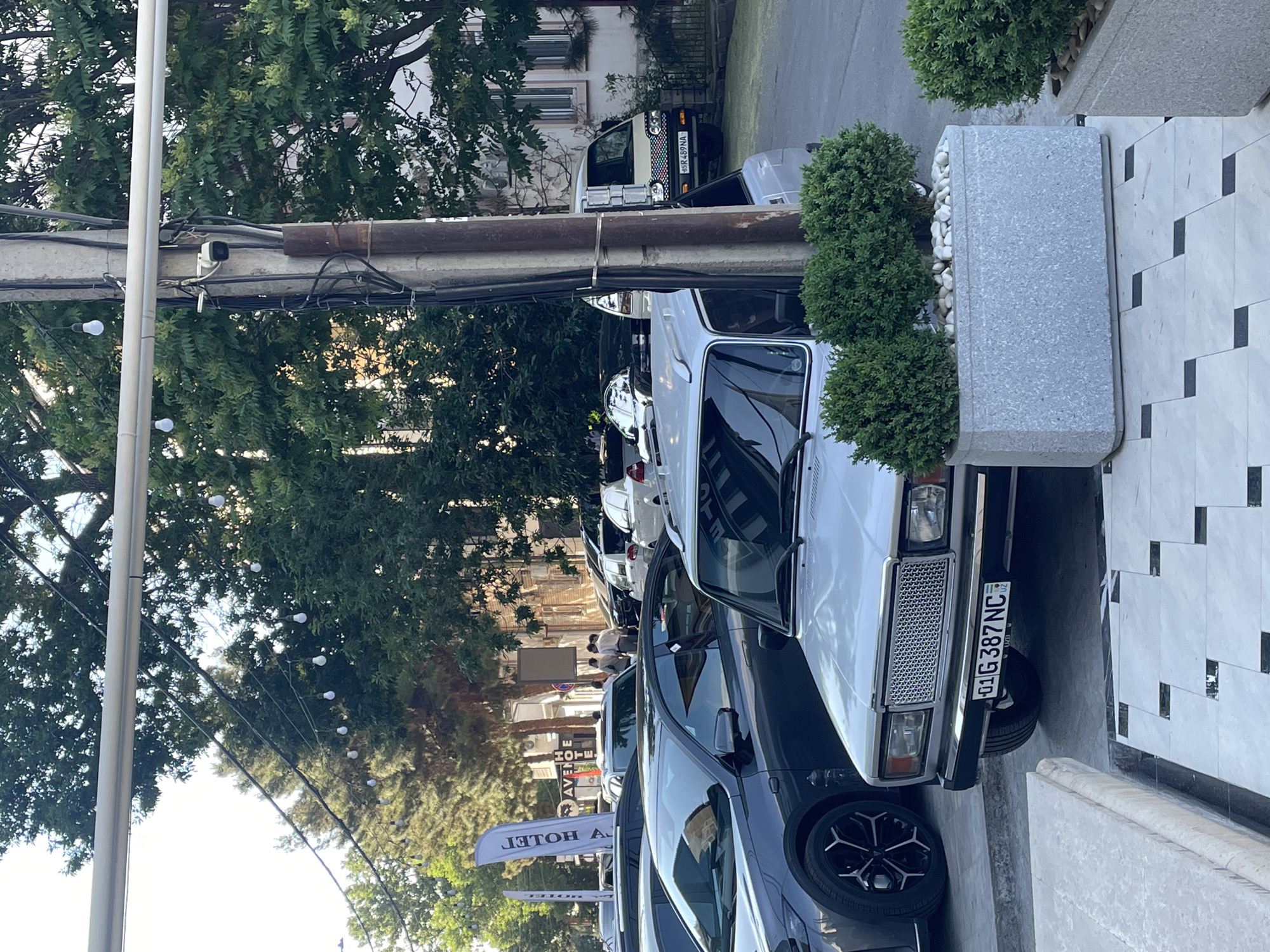 The image shows a street scene in Tashkent, Uzbekistan. Several cars are parked along the road, including a white Lada with license plate "01 G 387 NC". A dark-colored car with black alloy rims is partially visible in the foreground. Trees line the street, and decorative lights are strung along a cable. A utility pole is present. A gray stone planter with bushes is beside the white Lada. People are walking on the sidewalk further down the street. A truck is visible in the distance. A sign for "Avenue Hotel" is visible.