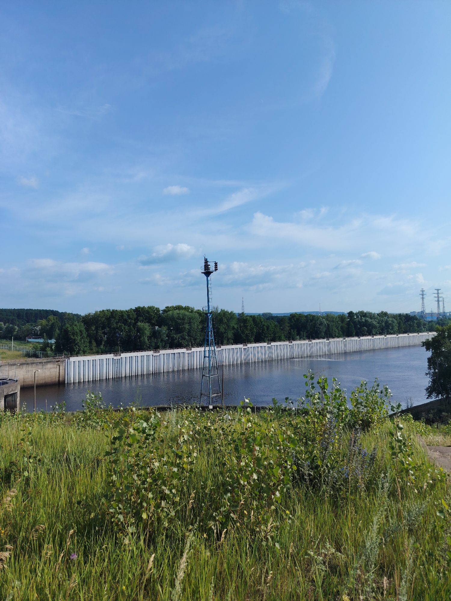 A wide shot depicts a dam on a river under a partly cloudy blue sky. The dam is a long, white, reinforced concrete structure with vertical pilings. A body of dark water extends from the dam towards the viewer. In the foreground, lush green grass and small bushes grow on a gentle slope. A blue metal structure with lights is positioned on the right side of the dam. In the distance, a dense line of green trees stretches across the horizon, with power lines and buildings visible beyond them to the right. The scene is likely the Votkinsk Dam in Chaikovsky, Russia.