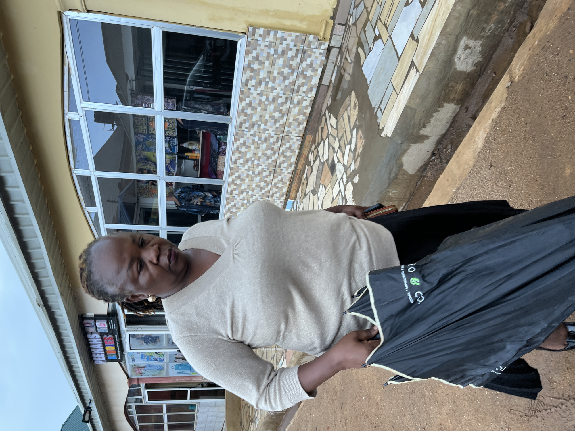A middle-aged Black woman is standing on a sidewalk next to a building. She is wearing a long-sleeved beige shirt and a black skirt or pants. She is holding a black umbrella in her right hand.
The building behind her has a storefront with glass windows. A sign above the storefront reads "Peenah's Touch". The tiles on the building are multi-colored and mosaic-like. The sidewalk is made of brown dirt.
The location is likely in Jos, Nigeria, based on the information provided.