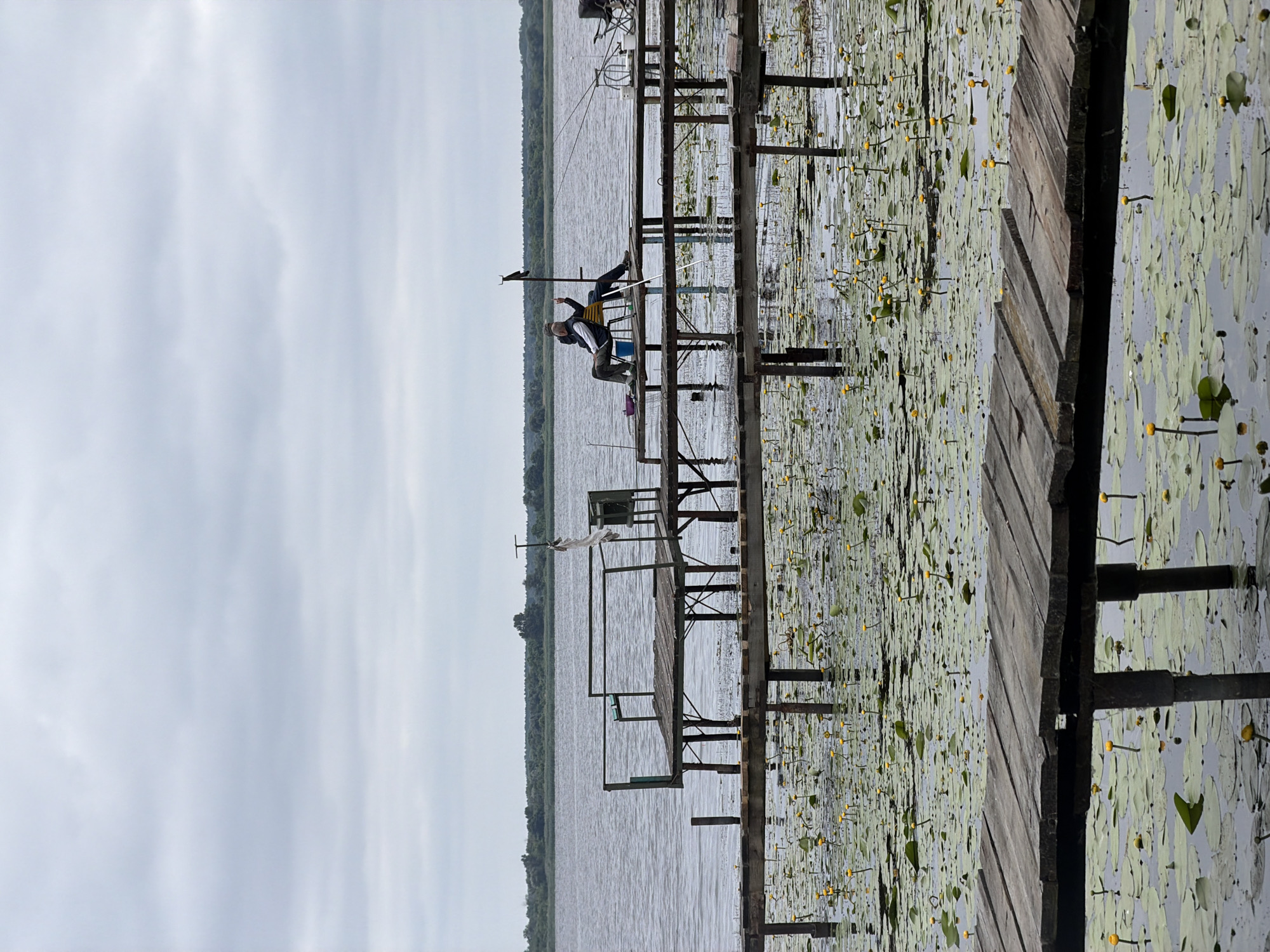 This is a photograph of a cloudy day scene at a lake in an unknown location. In the foreground, a wooden pier extends out over the water, which is covered with numerous lily pads and small yellow water lilies. Another, larger wooden pier is visible in the mid-ground, also over the lily pad-covered water. On this pier, two individuals are seated on chairs, appearing to be fishing. One person is looking towards the water, while the other is gesturing with their left hand. Fishing rods are set up on the pier. In the background, a tree line is visible across the lake. The sky is overcast with a significant amount of cloud cover.