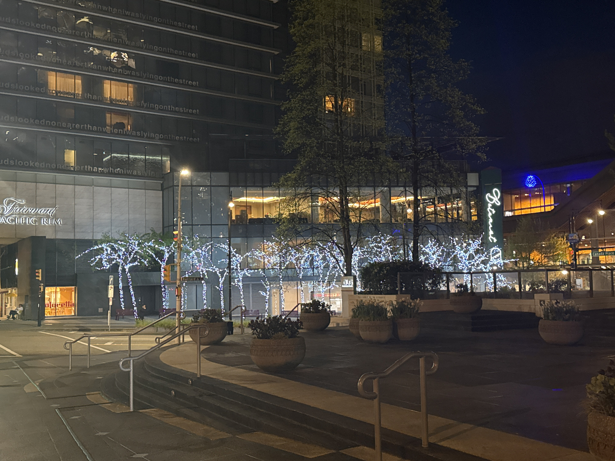 The image depicts a nighttime scene in Vancouver, Canada, specifically the area surrounding the Fairmont Pacific Rim hotel and the Burrard Skytrain station. The foreground features a multi-tiered pedestrian area with planters and metal railings. In the background, the Fairmont Pacific Rim building is visible, with illuminated windows and text art on its facade. Trees decorated with white lights line the street. To the right, a portion of the Burrard station is visible, along with signage for Botanist restaurant and Canada Place in the distance.