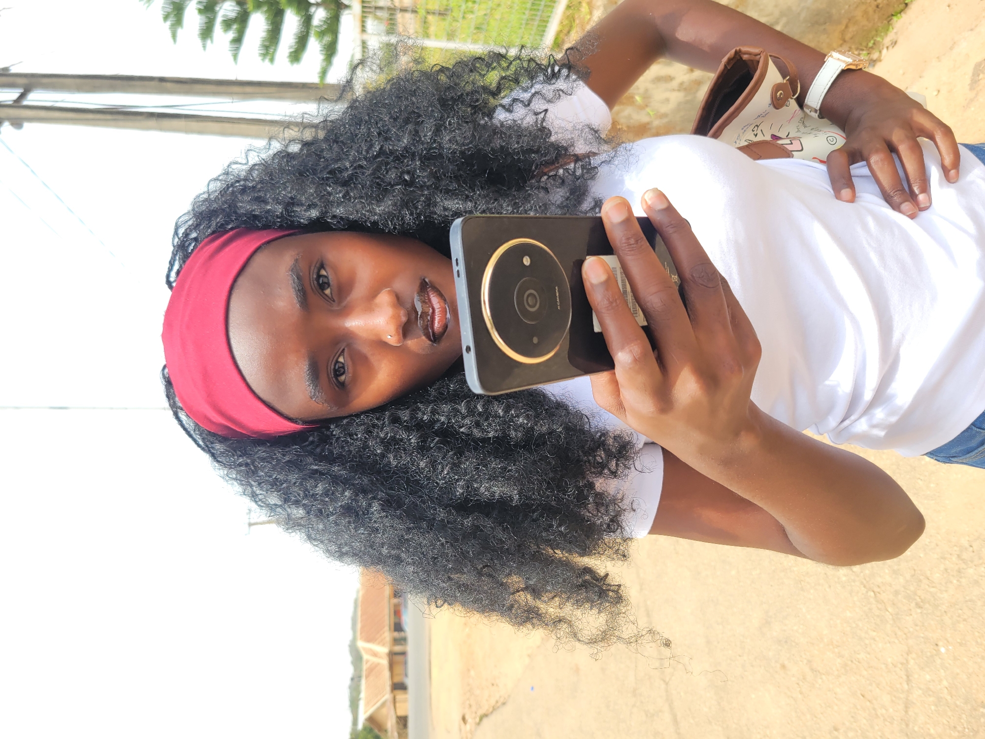 A person with dark skin is holding a smartphone in their right hand and pointing it at the camera, taking a selfie. The phone has a large circular camera module with a gold ring around it. The person is wearing a white t-shirt and a red headband. They have dark, curly hair. On their left wrist is a white wristwatch with a gold face. Behind the person is a light-colored, possibly beige, surface. The location is Jos, Nigeria. A pole and greenery are visible in the background. A building appears in the distance.