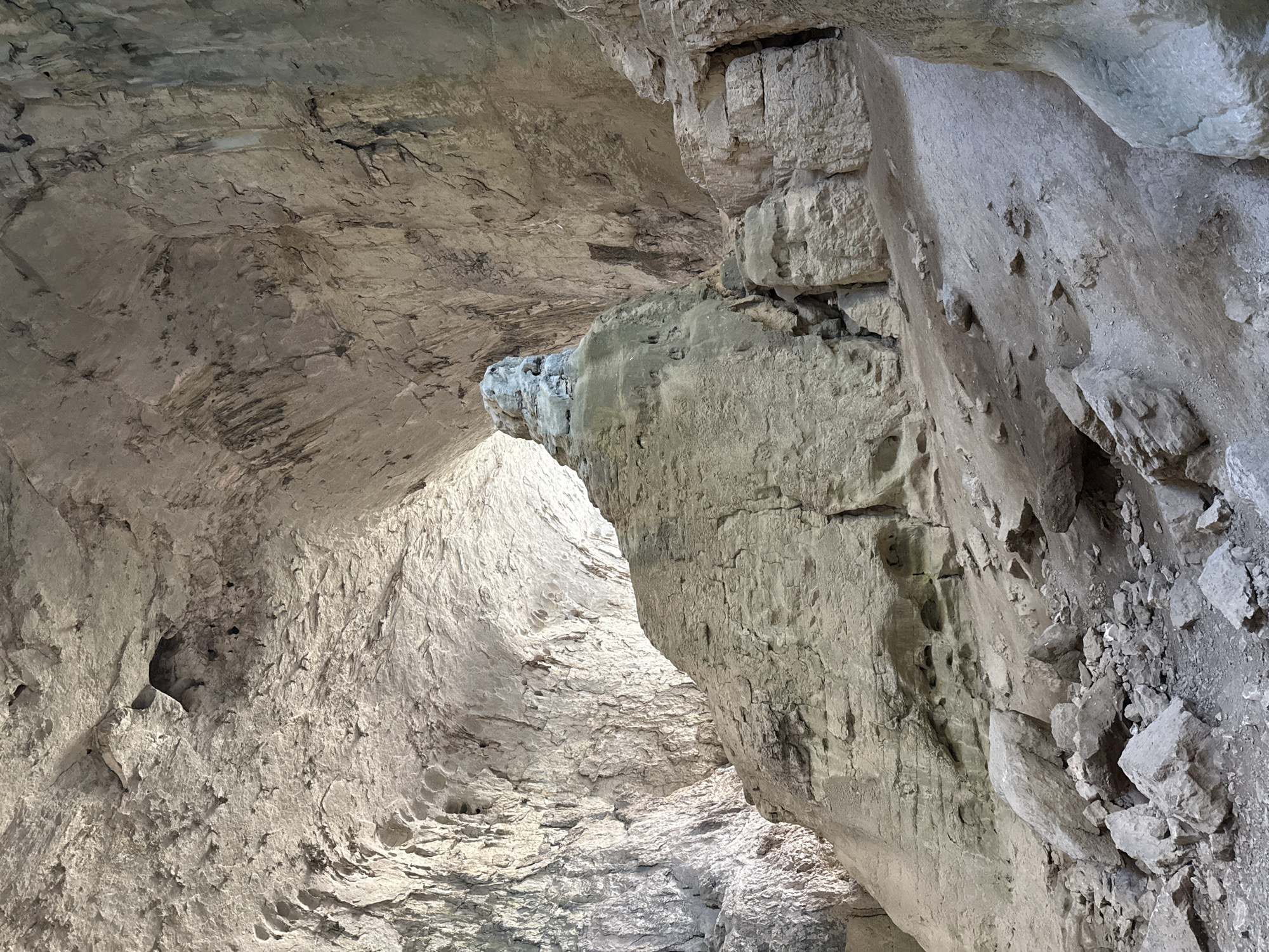 The image displays the interior of a cave, likely in Karlukovo, Bulgaria, characterized by rough, textured rock formations and a high, arched ceiling. The lighting suggests daylight entering from an opening, illuminating the cavern and casting shadows. Dominating the center is a large, irregularly shaped rock pillar, which appears to be a significant geological feature within the cave. The ground is covered in loose rocks and dirt, indicating a natural, undeveloped environment. The cave walls exhibit layering and erosion, suggesting a long geological history. The overall impression is one of natural grandeur and geological timescale.