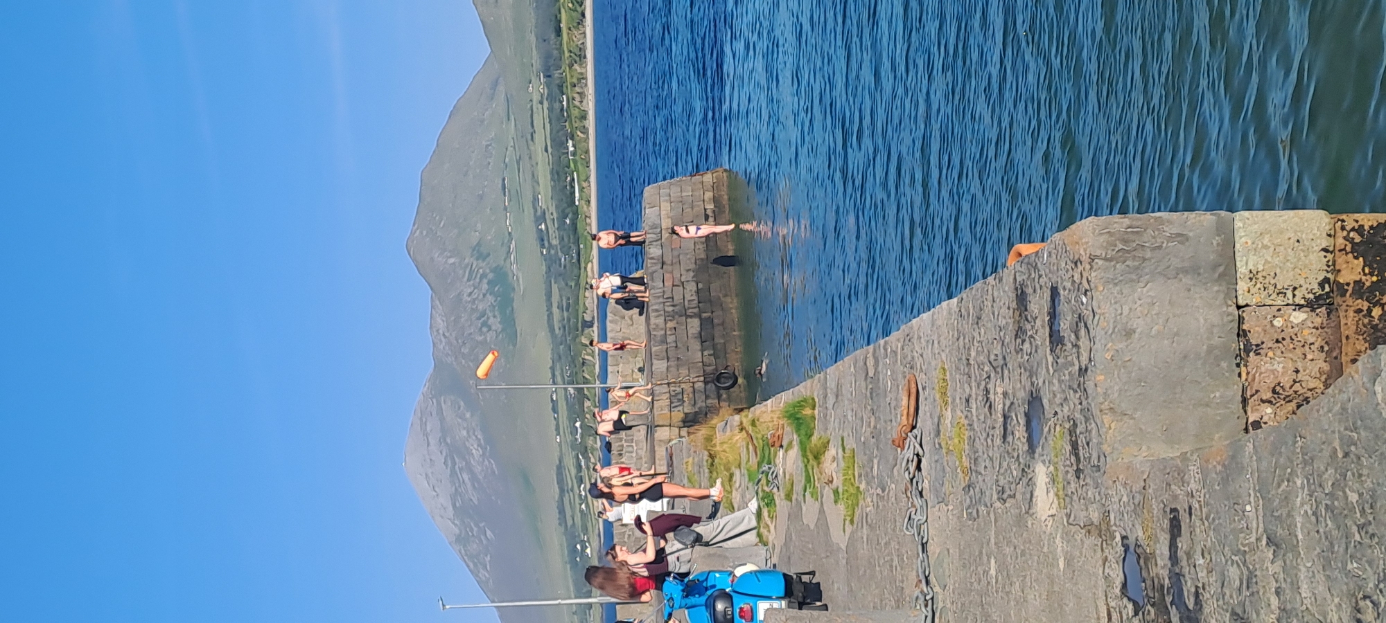 The image depicts a sunny day at a stone pier in an unspecified location in Ireland. A group of people are gathered on the pier, with some standing and others appearing to be preparing to jump into the bright blue water. One individual is visible in the water, having already jumped. In the background, a large, rugged mountain range dominates the landscape, with a few scattered houses and green fields visible on its lower slopes. An orange windsock is prominently displayed on a pole near the edge of the pier. The pier itself is constructed of large, weathered stones, with a thick chain and a tire hanging from it. A blue scooter is parked on the left side of the pier. The overall scene suggests a popular spot for recreational activities on a clear day.