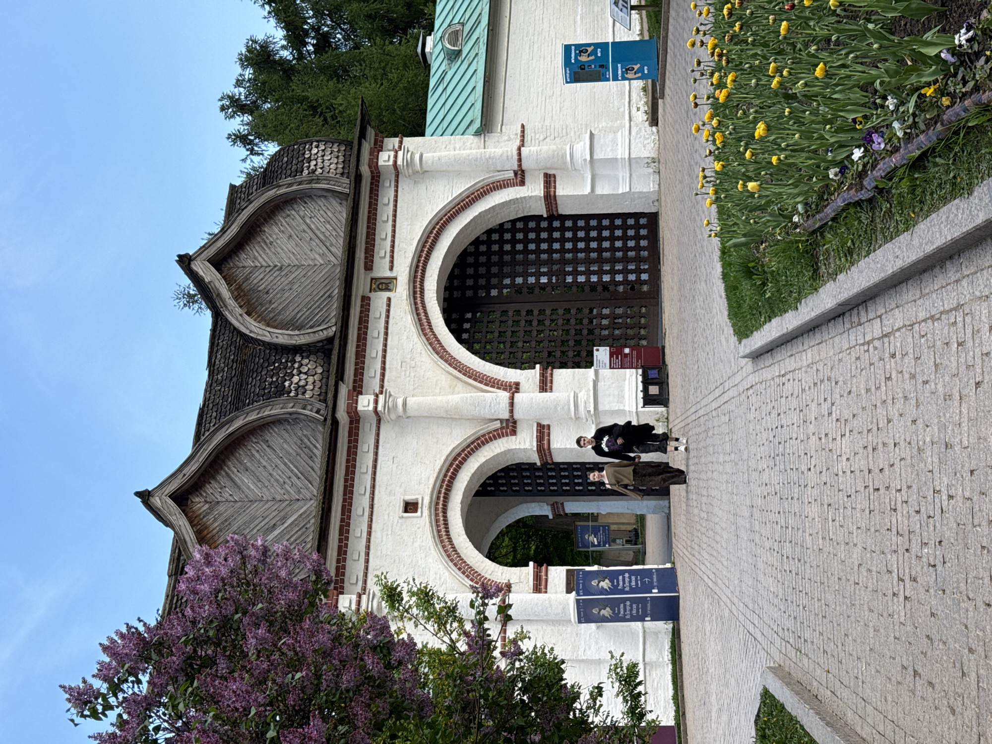 The image depicts the front entrance of the Church of the Ascension in Kolomenskoye Park, Moscow, Russia. The entrance features a white facade with two arched openings, wooden doors with a lattice pattern, and a unique layered wooden roof design. Two people are walking toward the entrance, a pathway paved with gray stones, flowerbeds with yellow tulips are on one side. Lilac bushes are also visible on the side of the entrance. Informational signs are posted near the entrance. The sky is visible above the building and the trees.