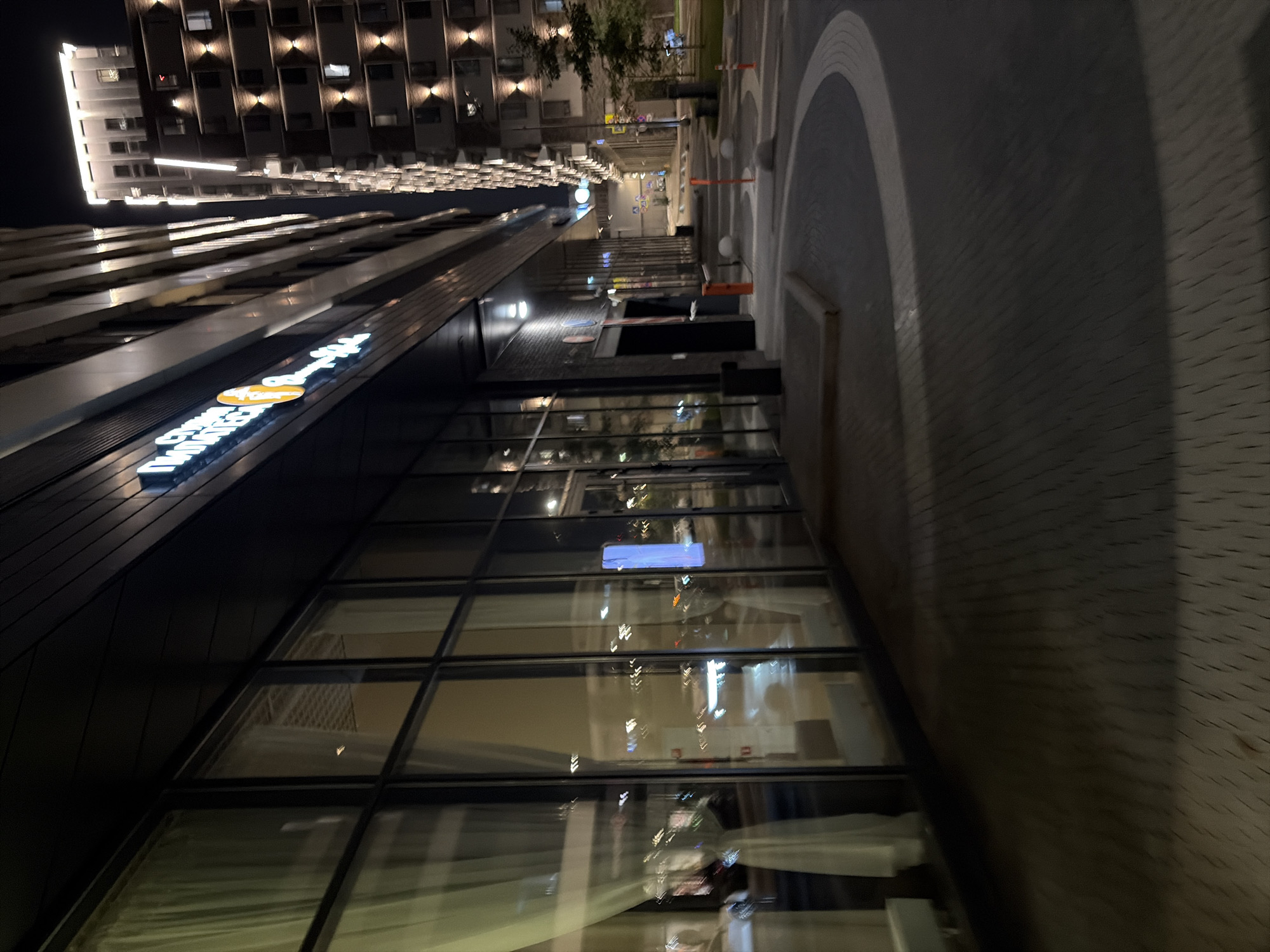 The image depicts a nighttime view of a modern commercial building with large glass windows. A neon sign with Cyrillic lettering is visible on the building's facade. In the background, another tall residential building stands illuminated. The street in front of the commercial building is paved with interlocking bricks and has a curved design. There are traffic bollards and street lights visible, indicating an urban environment. The reflection of lights and possibly vehicles can be seen in the glass windows. The overall scene suggests a well-lit, contemporary urban setting in Moscow, Russia.
