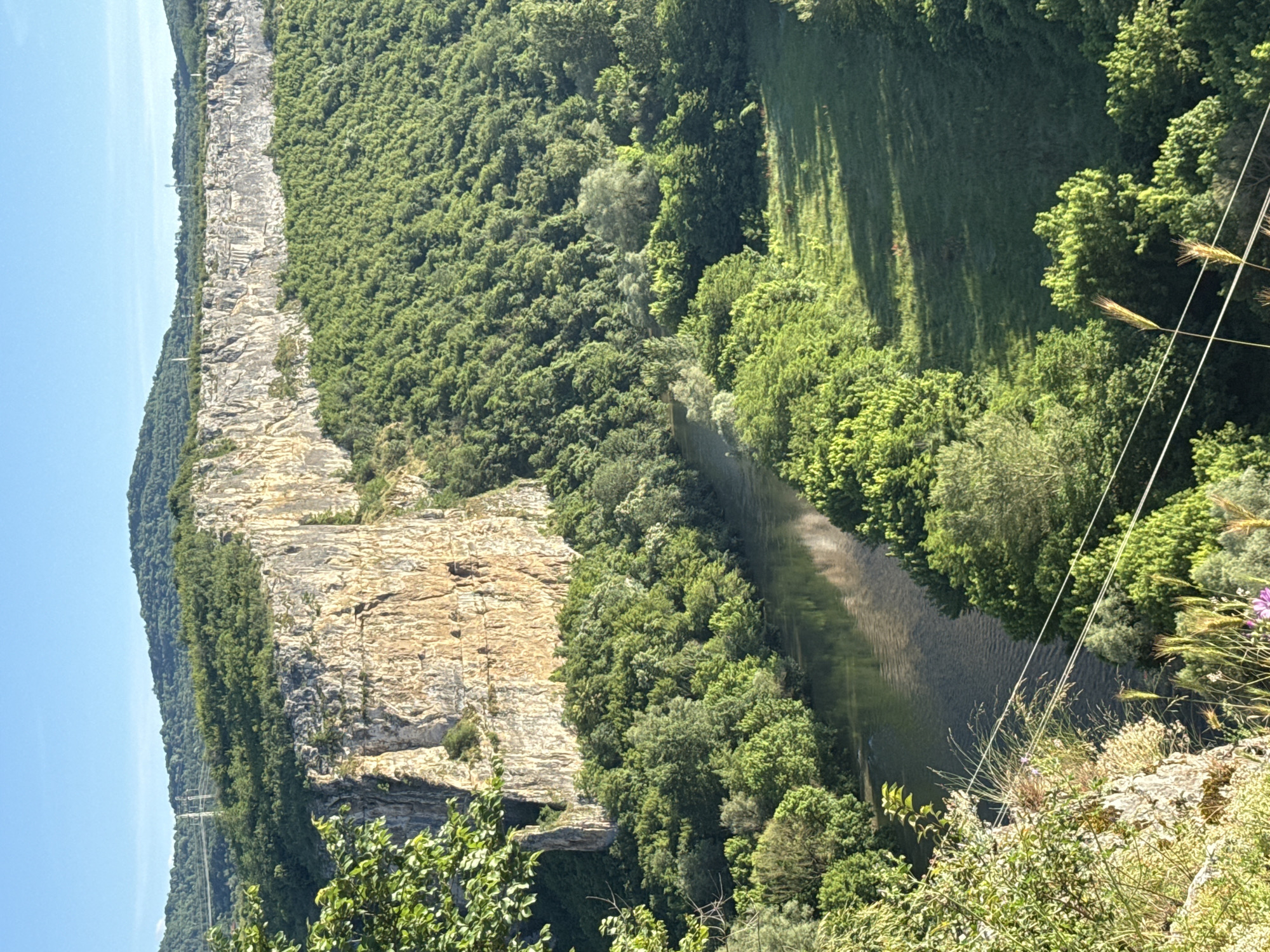 This is a high-angle, full outdoor shot of the Karlukovo Karst region in Bulgaria, featuring the Iskar River. A sheer, light-colored rock cliff face dominates the left side of the image, with its textured surface catching the sunlight. Dense, vibrant green foliage covers the slopes of the hills and extends along the riverbanks, creating a lush, natural landscape. The Iskar River flows gently through the scene, its surface reflecting the sky and surrounding trees. In the distance, rolling hills covered in trees form the horizon under a clear blue sky. Power lines are visible traversing the landscape in the upper left and lower right portions of the frame. There are no people or animals depicted in the image.