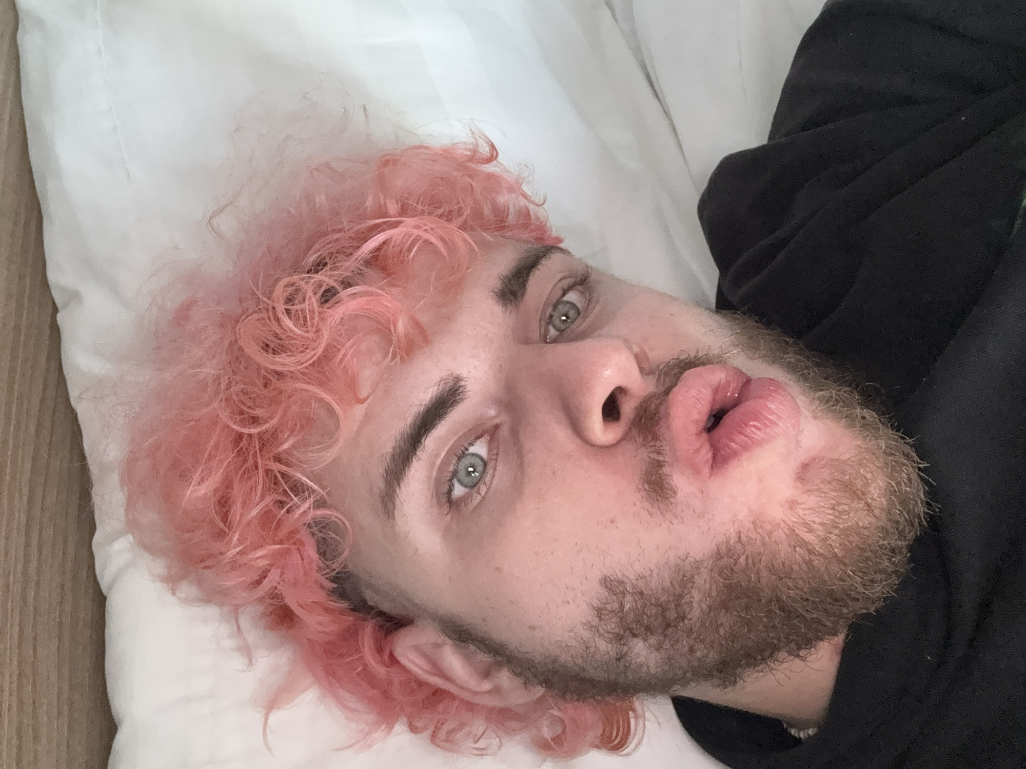 A man lies on his back on a white pillow. He has pink curly hair, a light beard, and is wearing a black sweatshirt. His face is tilted slightly upward, and his lips are slightly puckered. The image appears to have been taken at a close angle. There is no indication of the location being in Đà Nẵng, Vietnam.