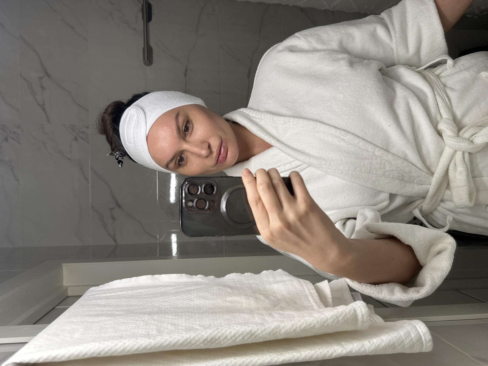 A woman in a white, terrycloth bathrobe and a white spa headband takes a mirror selfie. Her dark hair is pulled back into a bun, partially visible behind the headband. She holds a dark-colored smartphone, equipped with a prominent triple-lens camera system and a circular MagSafe accessory, positioned to capture her reflection. Her expression is neutral and calm, with her lips slightly parted.
The setting appears to be a modern bathroom or spa area, characterized by light grey and white marble-patterned wall tiles. A metallic fixture, possibly a shower head, is reflected in the upper left background. Below the mirror, a stack of neatly folded white towels rests on a white ledge or surface. The overall impression is one of relaxation and self-care, possibly before or after a shower. While the image does not provide explicit location cues, the user indicates the scene is in Moscow, Russia. Partially visible text on a tag inside the bathrobe sleeve appears to read "ALCОBA".