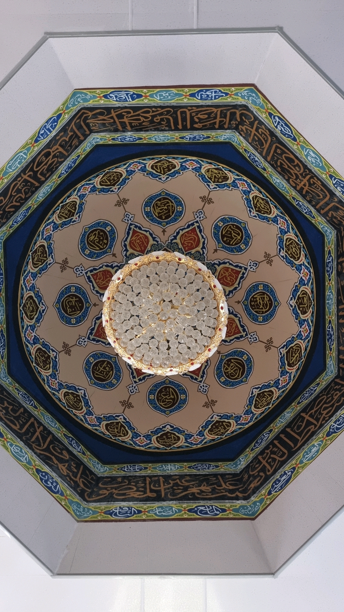 This is a high-angle, full shot of the interior ceiling of a mosque in Lhoksukon, Indonesia, featuring an ornate chandelier. The ceiling is octagonal, with intricate Islamic calligraphy adorning its surfaces. The calligraphy is predominantly in brown and blue hues, set against a beige background, and framed by geometric patterns in blue, green, and red. A multi-faceted crystal chandelier, with a gold-colored frame, hangs at the center of the ceiling. The chandelier is the focal point, reflecting light and adding a sense of grandeur to the space. The surrounding walls are plain and white, providing a contrast to the richly decorated ceiling. The image appears to have been taken from a slightly tilted angle, giving a dynamic perspective of the ceiling's architecture and design.