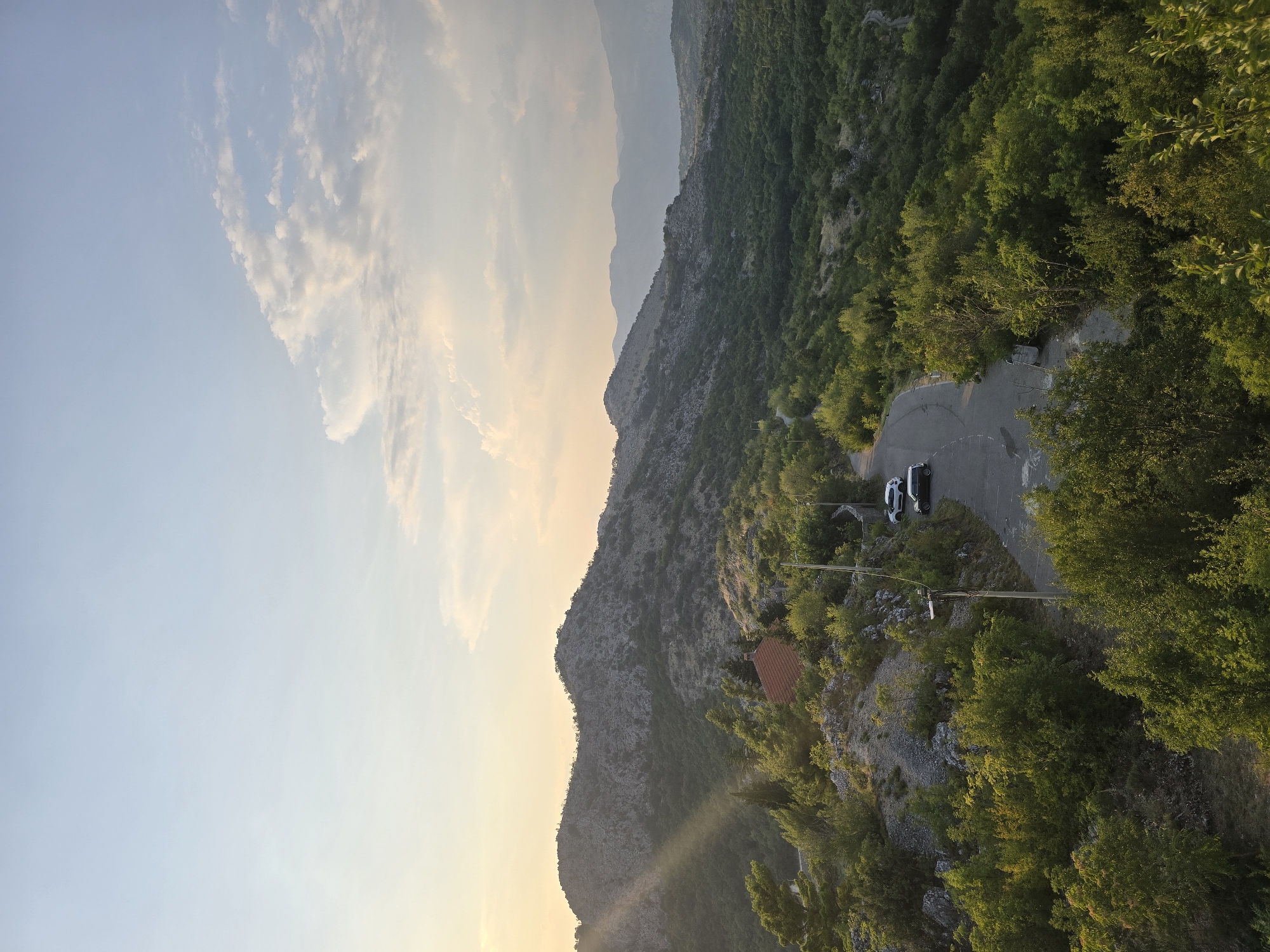 The image depicts a scenic landscape in the vicinity of Gornja Vrbica, Montenegro. A winding asphalt road cuts through a verdant, hilly terrain dotted with trees and rocky outcrops. The sun is setting, casting a warm glow and creating visible sunbeams that slant across the left side of the frame, illuminating a small, reddish-roofed building nestled among the trees. Two cars, a white SUV and a dark sedan, are parked on the side of the road near the building. In the background, layered mountain ranges fade into the distance under a pale blue sky with scattered clouds. The overall scene conveys a sense of natural beauty and tranquility, characteristic of a rural mountainous region.
