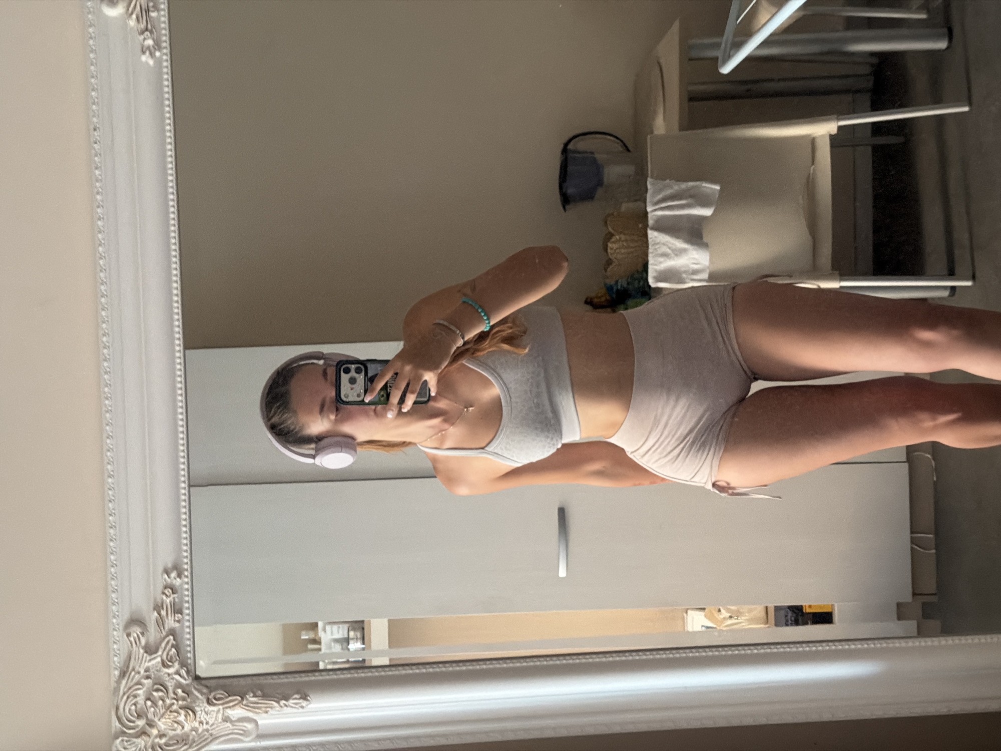 A young woman, wearing headphones and a gray sports bra with light pink shorts, takes a mirror selfie in what appears to be an apartment in Sofia, Bulgaria. She is holding a smartphone in her right hand, capturing her reflection. The background shows a large ornate mirror, a white wardrobe with a silver handle, a table with a water pitcher and a white towel, and two chairs. The lighting suggests it is daytime, with natural light entering the room.
