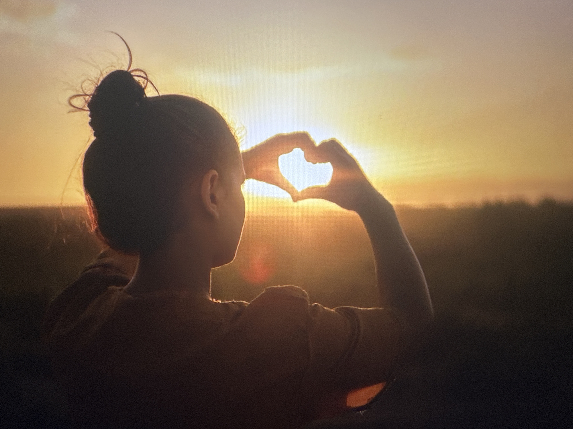 The image shows a silhouette of a person's head and shoulders against a sunset. The person has their hair in a bun and is looking towards the sunset. They are holding their hands up to form a heart shape, with the sun shining through the opening of the heart. The background is a hazy, golden sky with some trees or land visible in the distance. The scene appears to be outdoors, possibly in a park or open field. There is no specific indication of this location being in Bryansk, Russia, or any other city.