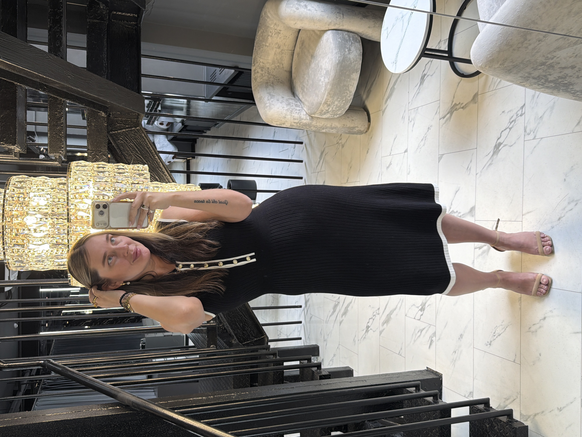 A woman is taking a selfie indoors. She is standing on the stairs of a black metal staircase, next to a chandelier. The floor is white marble. In the background is a white armchair and a small marble table. It is not possible to determine whether this is the city of Znamya Oktyabrya, Russia.