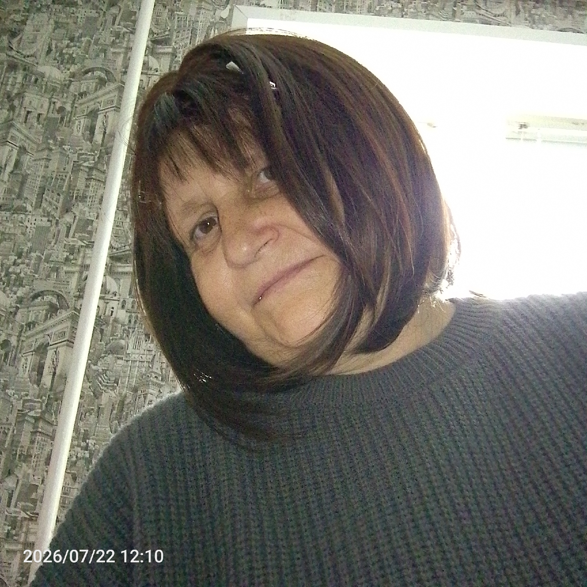A medium shot captures a woman from the shoulders up, indoors. She has brown hair with bangs and is wearing a dark gray sweater. The background features a patterned wallpaper with what appears to be a cityscape design, and a white vertical object, possibly a pipe or curtain rod, runs down the left side of the frame. To the right, a bright area suggests a window or light source. A timestamp "2026/07/22 12:10" is visible in the lower left corner. The woman's face is tilted slightly upwards, with her eyes looking towards the camera. Her expression is neutral.