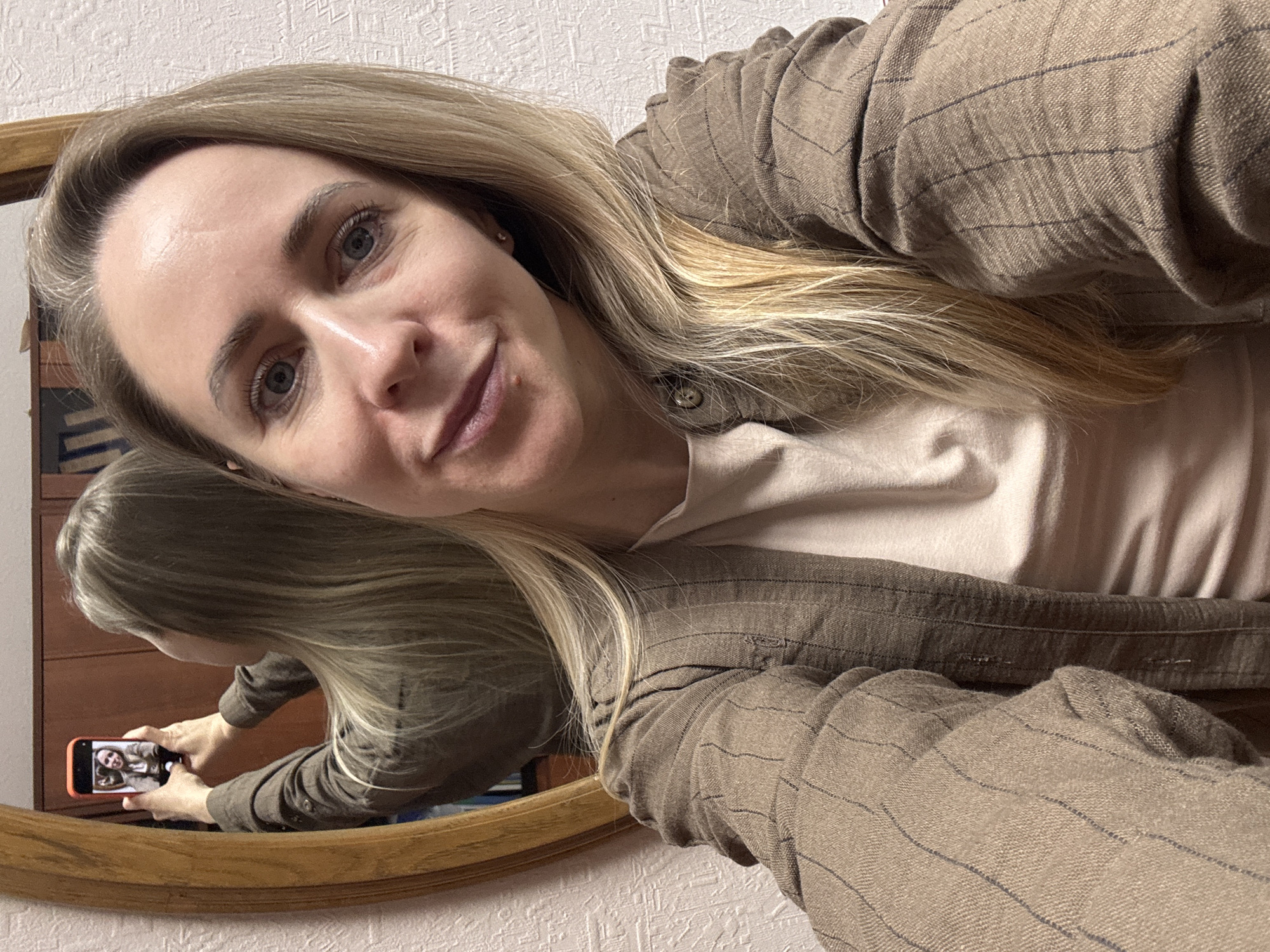 A woman with blonde hair is taking a selfie in a mirror. She is wearing a brown striped jacket over a light pink top. The reflection in the mirror shows her holding a phone, presumably taking the picture. The background appears to be a room with textured wallpaper. There is no information to indicate this is in the city of Chaikovsky, Russia.