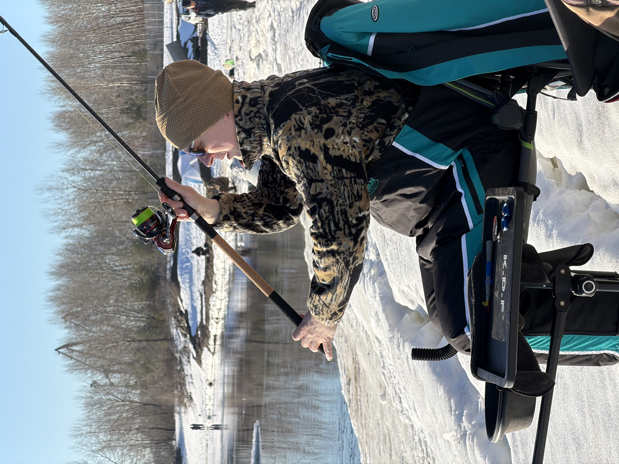 A person is ice fishing, dressed in a tan knit hat and a camouflage patterned jacket, standing near a body of water with snow and ice on the banks. The person is holding a fishing rod with a red and silver reel. To the person's right is a device with blue and black elements, potentially a specialized fishing stand or chair with accessories. The environment includes bare trees along the distant shore and a partially visible group of people in the background. The sky is clear and blue. The setting appears to be a winter scene on a frozen body of water. The location is Noviy, Russia.