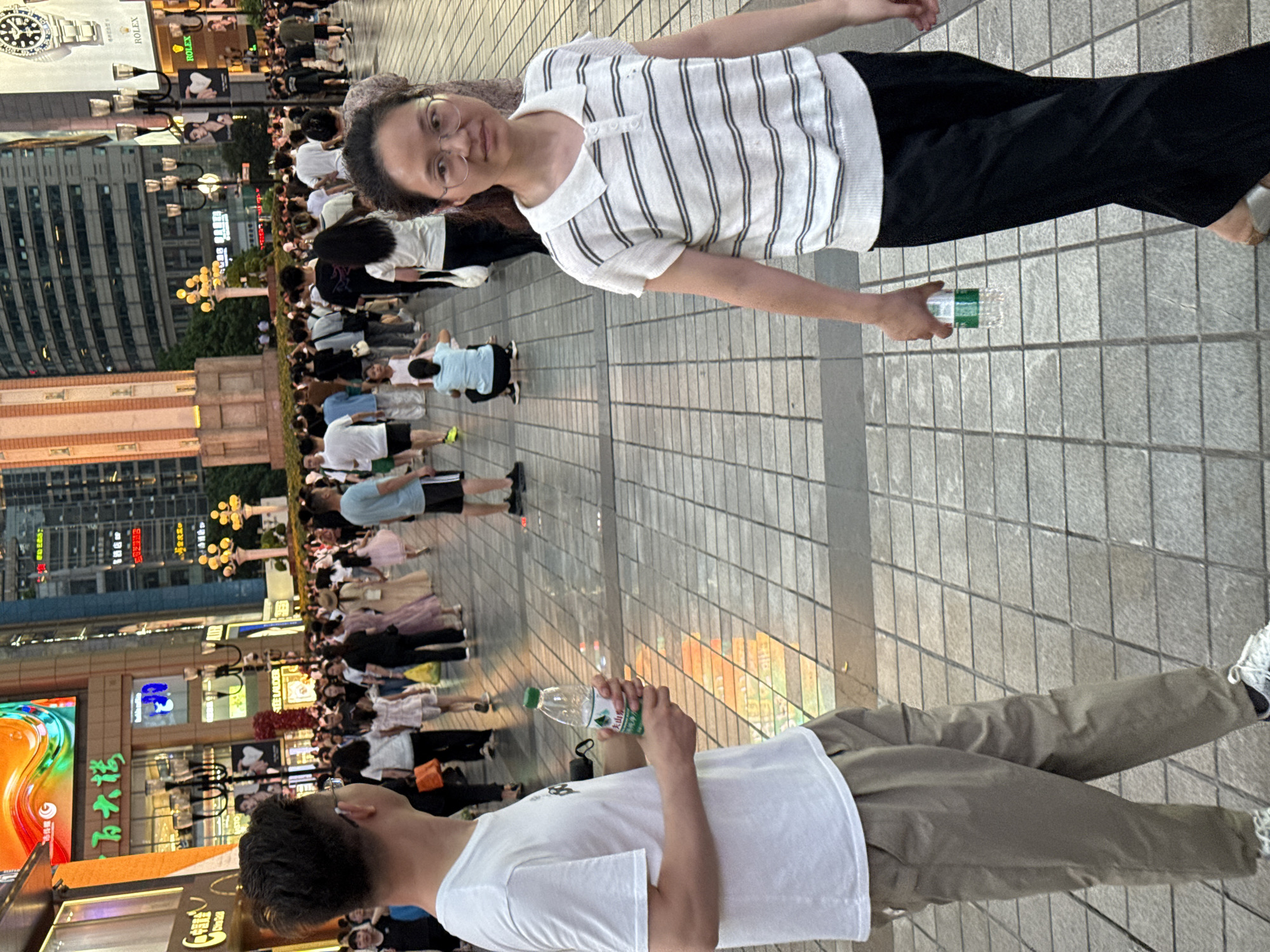 In the Yuzhong District of China, a street scene unfolds with numerous people walking and standing on a paved walkway lined with shops and buildings. The lighting suggests it is late afternoon or early evening.
In the foreground, a man wearing a white t-shirt and khaki pants is shown from the waist down, holding a plastic bottle of water. To his right, a woman in a striped polo shirt and black pants is captured from the chest down, also holding a plastic bottle of water.
Further back, a crowd of people are visible, some walking, others standing. A child in a light blue shirt is crouched down in the middle ground. The background features multi-story buildings with illuminated signage, including what appears to be a Rolex advertisement on one building. The overall environment is a bustling urban street, likely a shopping area.