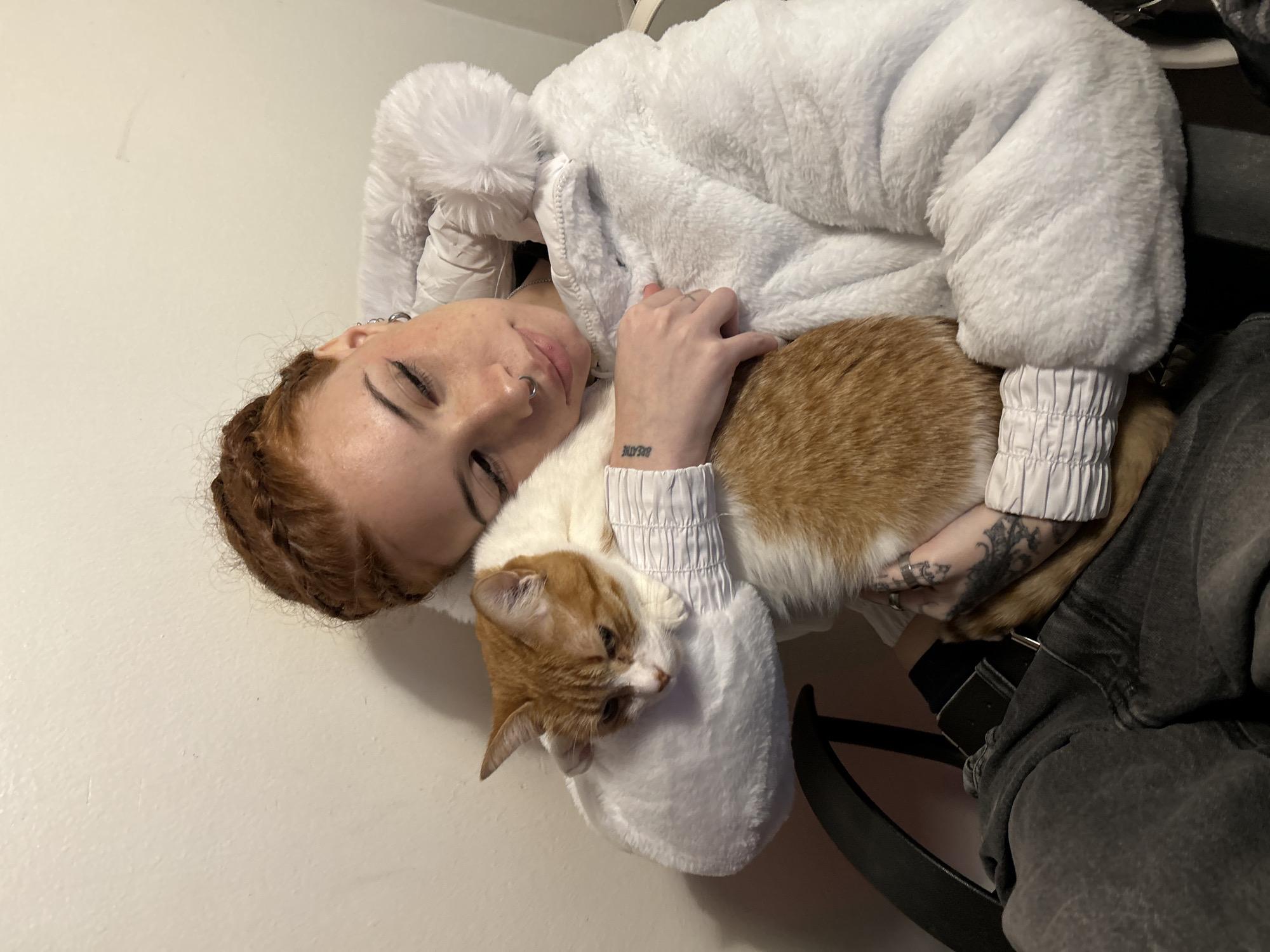 A pale-skinned, young adult female with red hair braided and pulled back from her forehead, lies on her side wearing a white, fluffy hooded jacket. She is holding an orange and white cat. The woman has her eyes closed and a nose ring. Tattoos are visible on her left forearm and hand. The cat is nestled against her and appears relaxed. The environment is a plain, light-colored wall in the background, and a chair visible on the right side of the frame, suggesting an indoor setting. The image is not taken in Buenos Aires, Argentina.