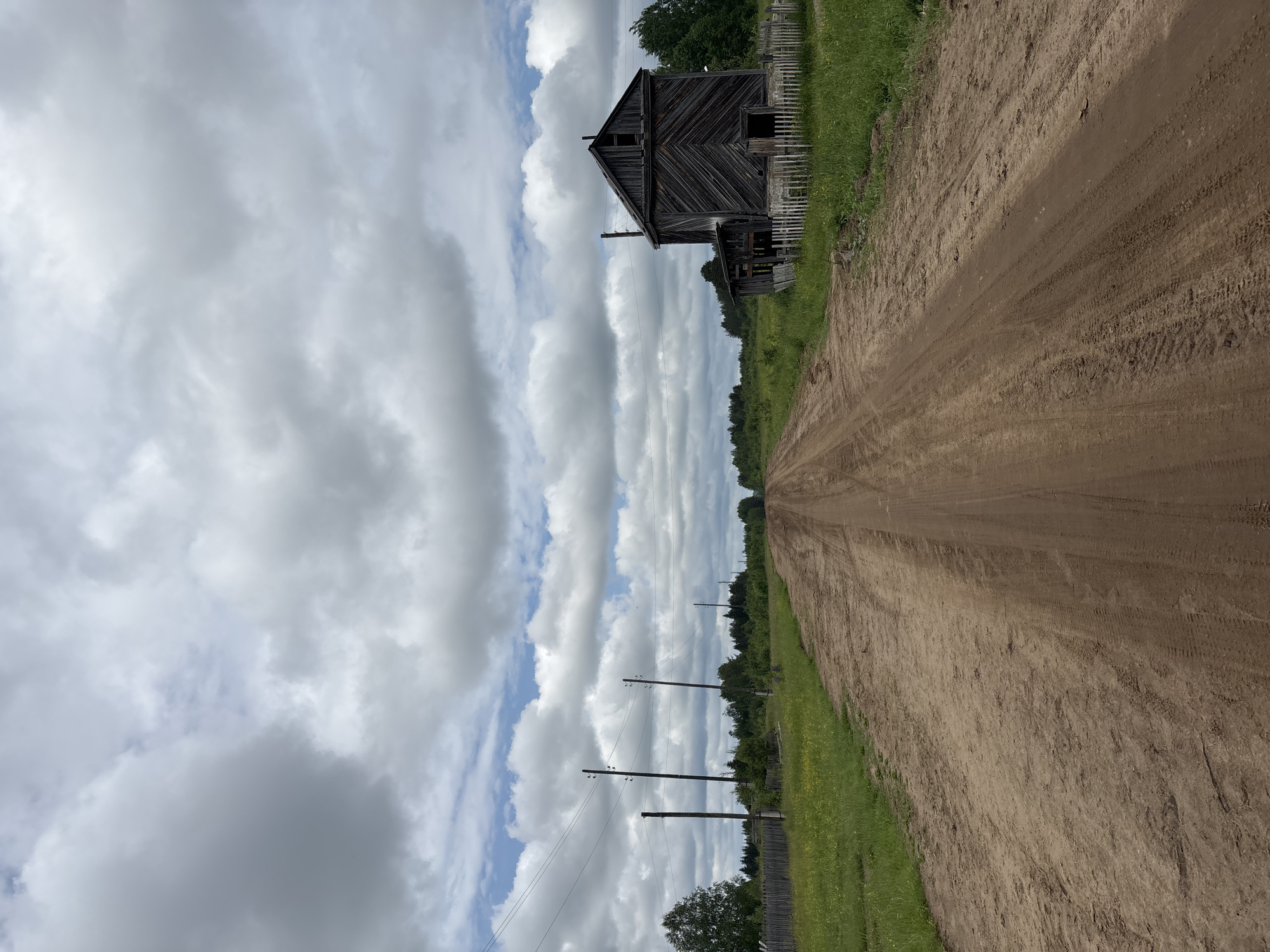 The image shows a dirt road leading into a rural area, likely in or near the city of Kurgan, Russia. The road is wide and appears to be unpaved, with visible tire tracks. To the right of the road is a weathered wooden building, appearing to be an abandoned barn or shed, with a small white picket fence in front of it. The building is dark wood and shows signs of disrepair, with missing planks and an open doorway. To the left of the road, there is a grassy field with scattered yellow flowers and a wooden fence. Further in the distance, there is a line of trees, and several utility poles with power lines are visible stretching across the landscape. The sky is filled with dramatic, cloudy formations, with patches of blue sky visible between them. The overall scene evokes a sense of remoteness and rustic simplicity. There are no people visible in the image.