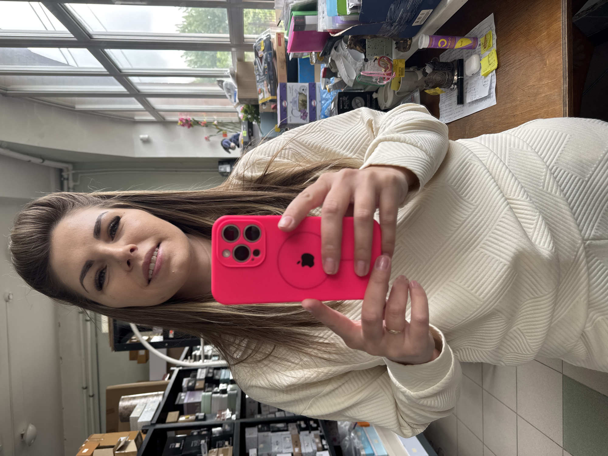 A woman holds a pink smartphone in her hands, possibly taking a selfie. The background includes what appears to be a cluttered workspace with various items on shelves and on a desk. It is not possible to determine the location as Klin, Russia, based on the given information.