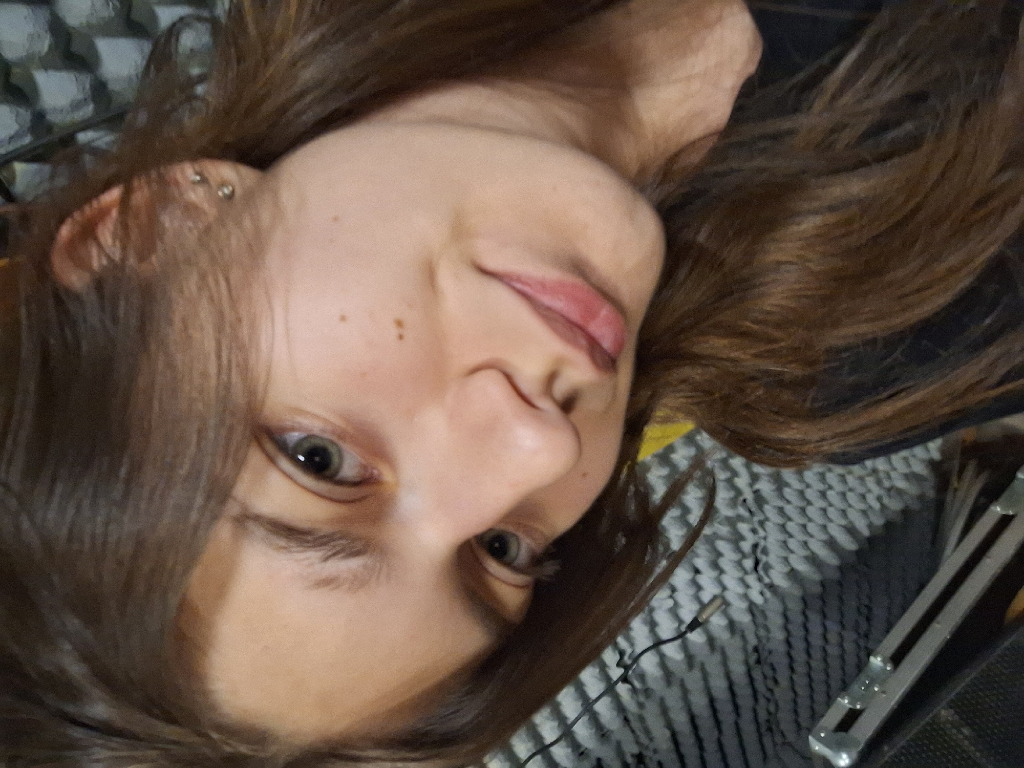 A close-up, angled portrait of a Caucasian woman with shoulder-length brown hair, who appears to be lying down. She has fair skin, light eyes, and a piercing on her left ear. Freckles are visible on her cheek. The background includes a gray and white patterned surface, possibly a blanket or shawl. An audio cable is also visible near her face. The woman's lips are slightly parted. No specific indicators pinpoint the location as Panagyurishte, Bulgaria. The description is based solely on the provided image.