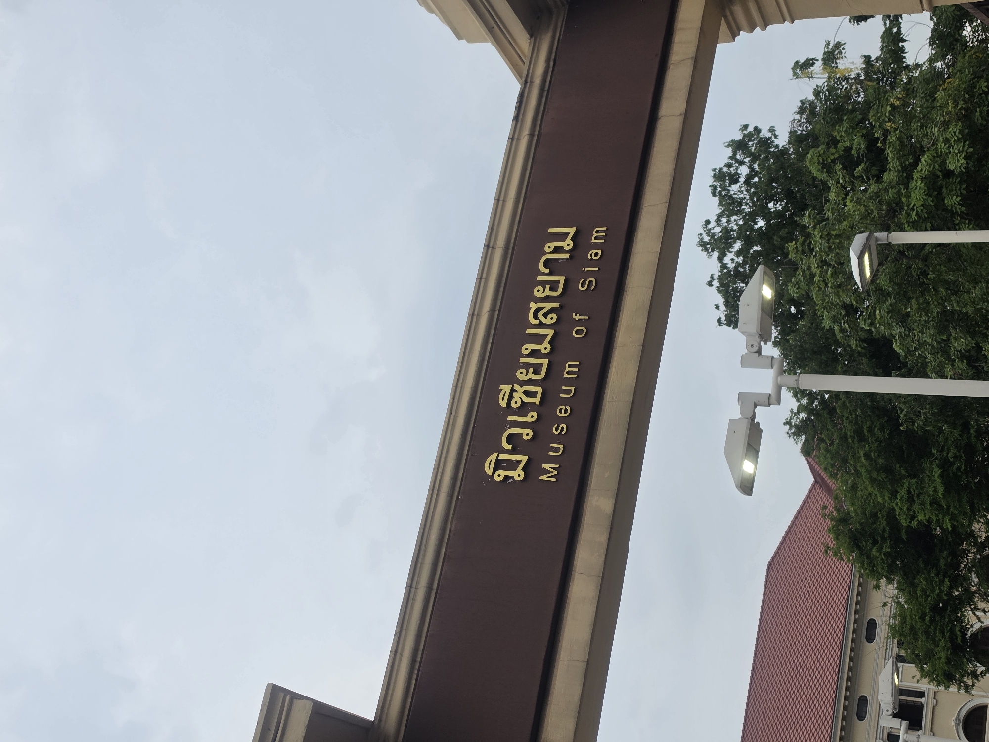 The image shows the entrance sign of the Museum of Siam in Bangkok, Thailand. The sign is a brown rectangular structure with the name of the museum written in gold letters, both in Thai and English. A streetlamp, trees, and a building with a red tile roof are visible in the background. The sky is overcast.